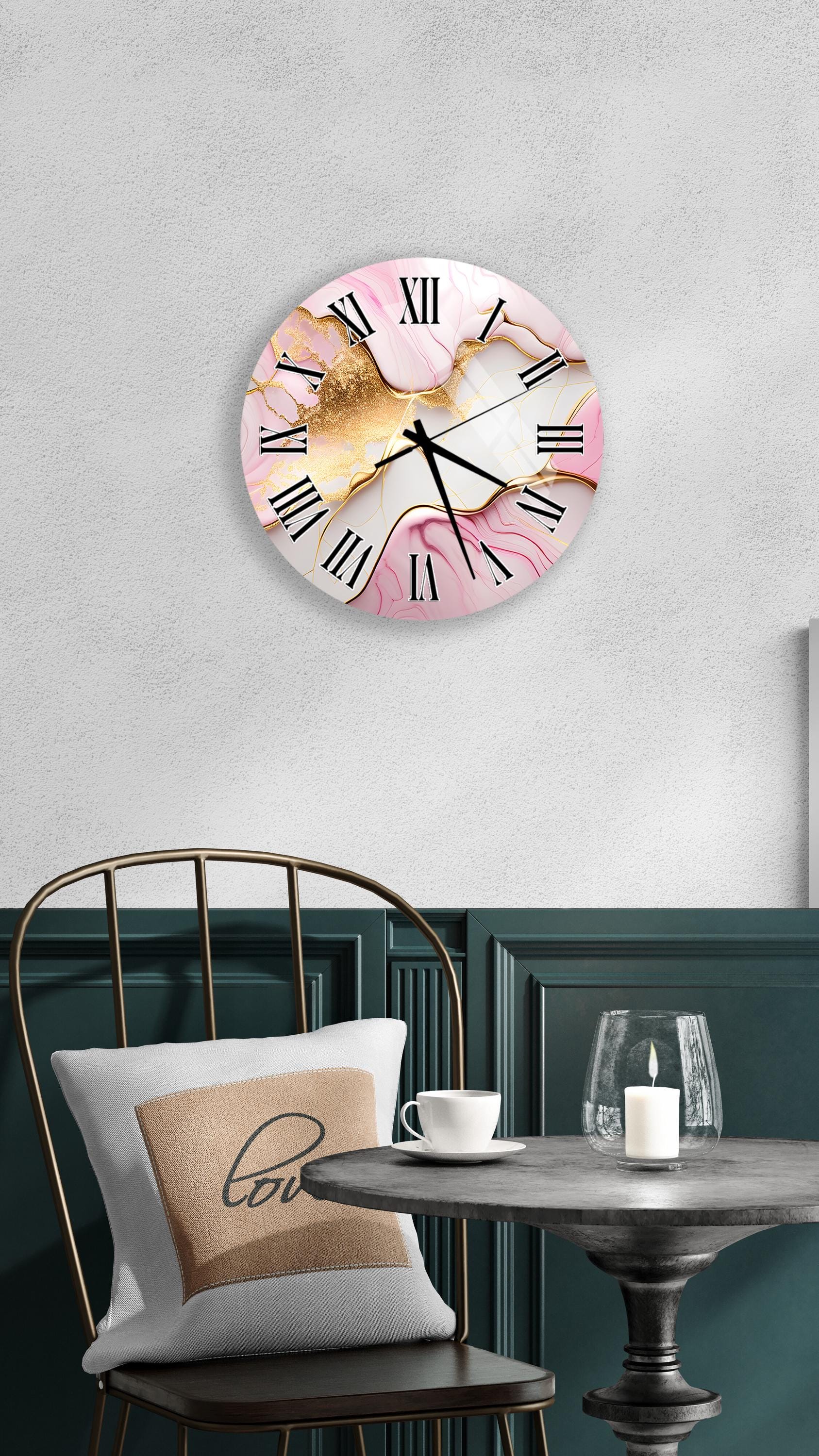 a wall clock with a pink and gold marble design, mounted on a wall above a table with a white tablecloth. The table has a glass candle and a white coffee cup on it. A white pillow with the word "love" printed on it is also present on the table.