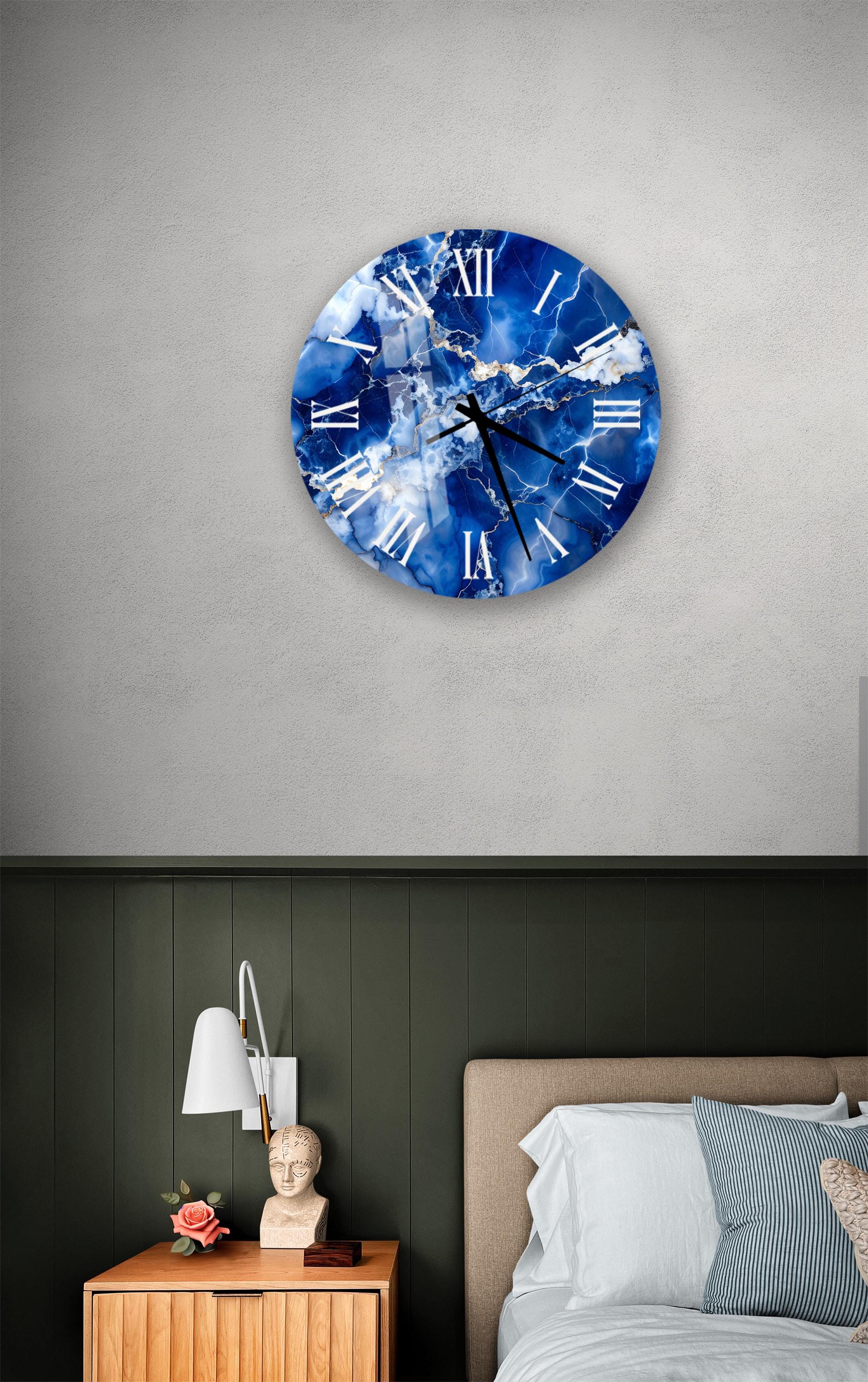 a bedroom setting with a large blue and white clock on the wall above a bed. The clock has a marble-like design and is mounted on a gray wall. The bed has a wooden headboard and is covered with white sheets. There is also a nightstand with a lamp and a small statue on it.