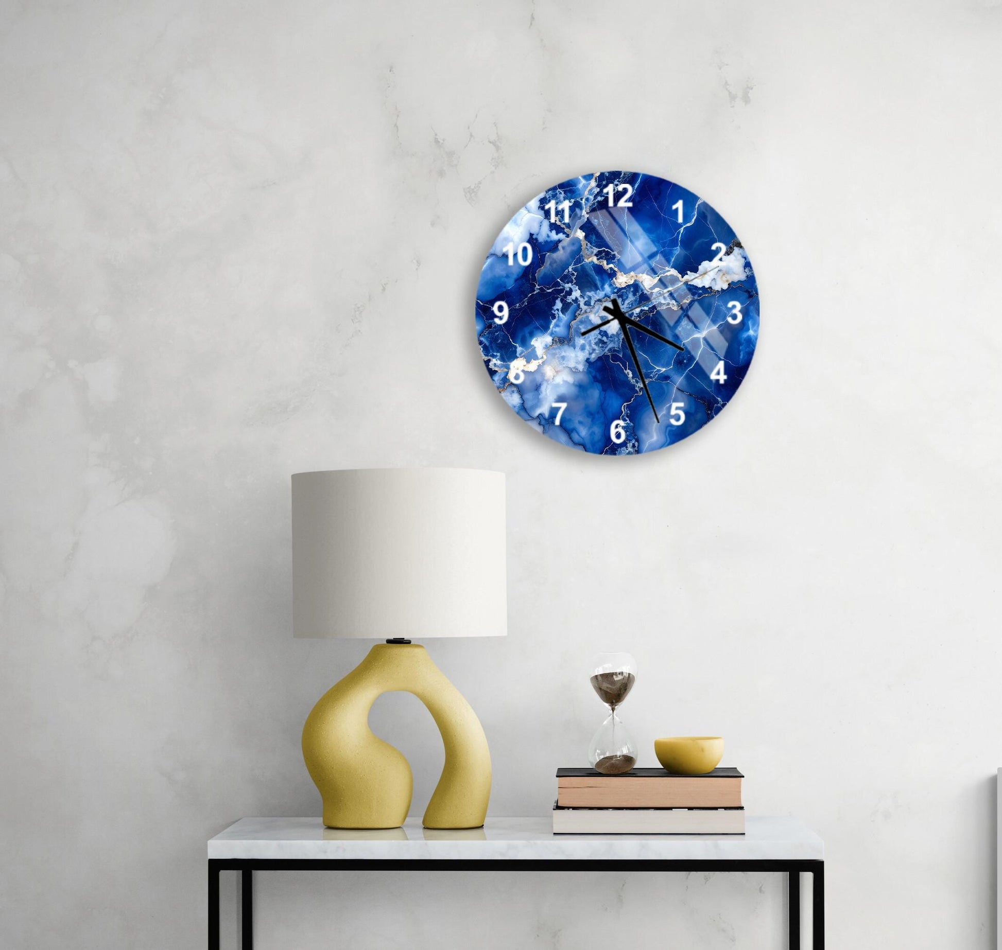 a modern wall clock with a blue and white design, mounted on a white wall above a white table with a yellow lamp and a stack of books.