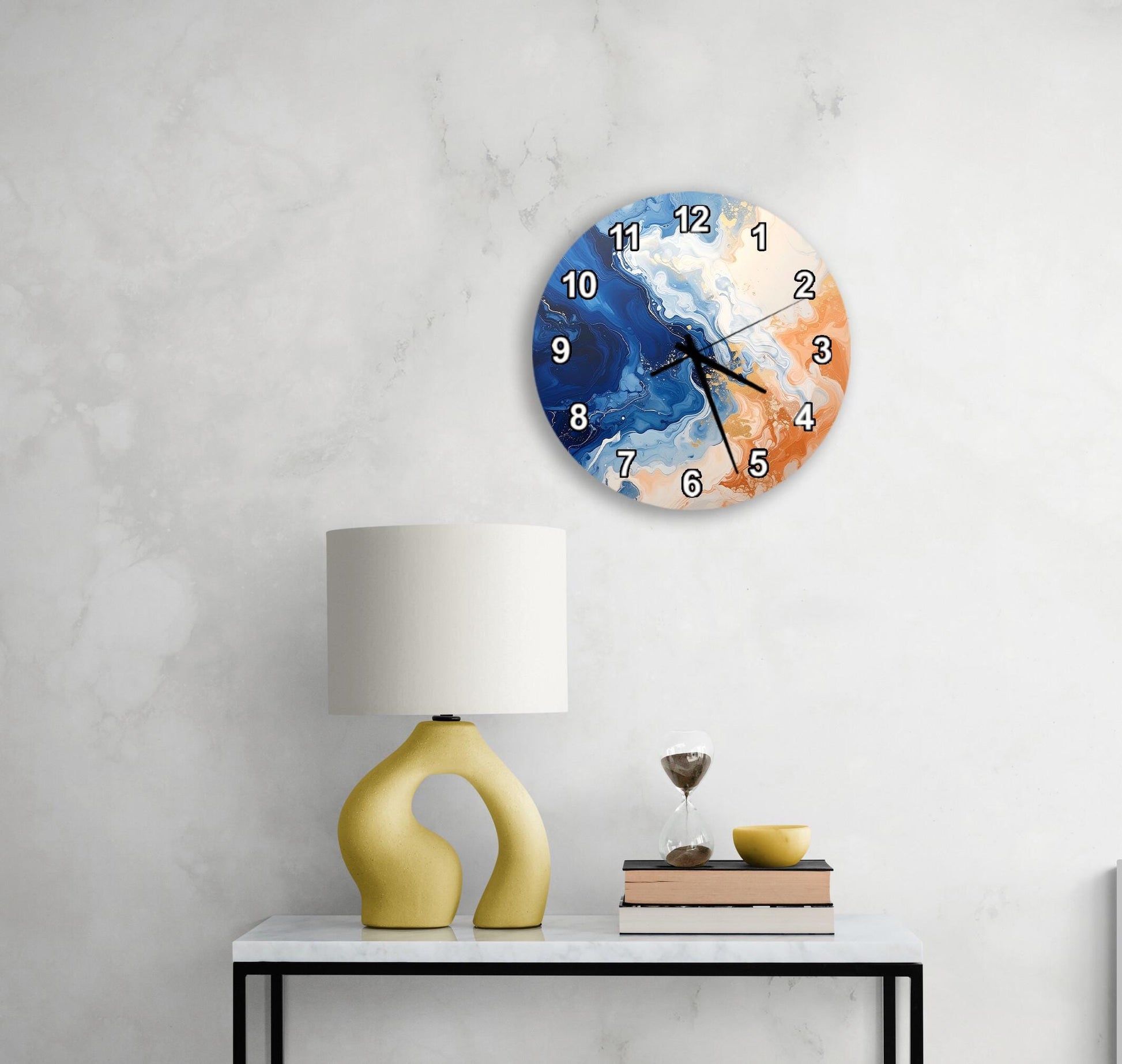 a modern wall clock with a blue and orange design, mounted on a wall above a white table with a yellow lamp and a stack of books.