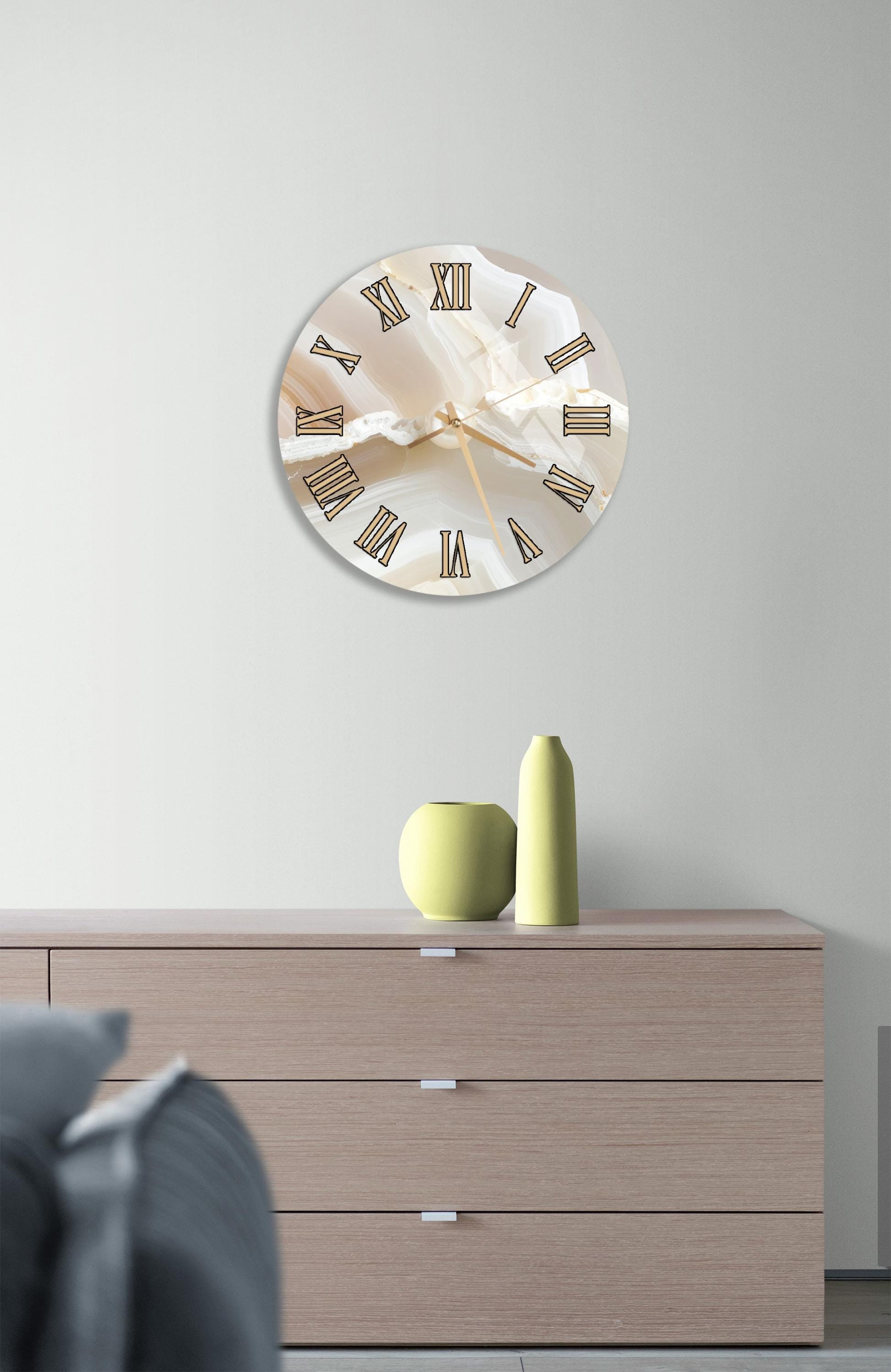 a modern living room with a large round clock on the wall above a wooden dresser. The clock has a white face with black Roman numerals and is positioned above a wooden dresser with three drawers. On the dresser, there are two small green vases and a yellow vase.