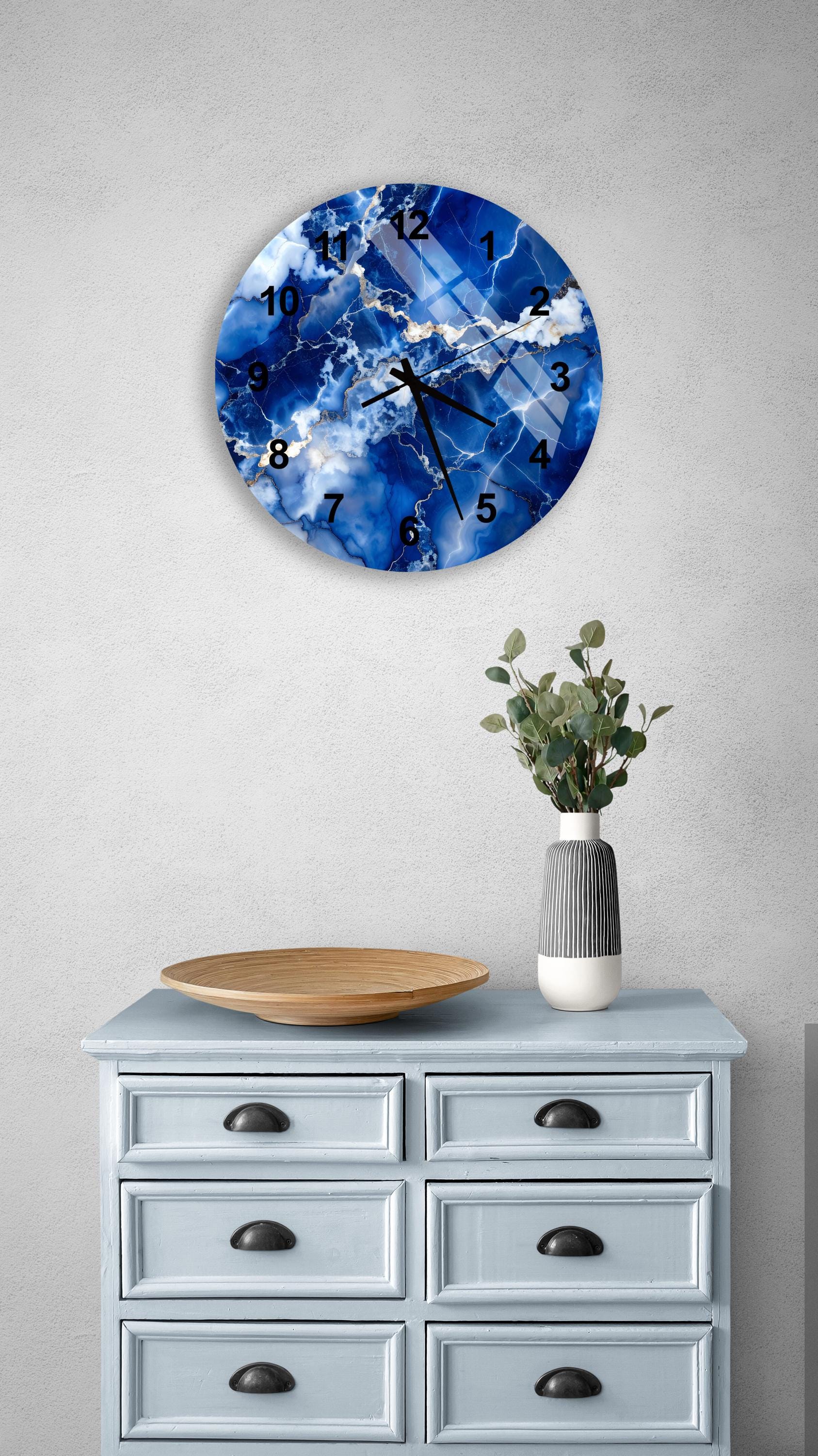 a blue and white marble clock mounted on a wall above a light blue dresser with six drawers. The clock has a marble-like design and displays the time. The dresser also has a vase with greenery on it, and there is a wooden bowl on top of it.