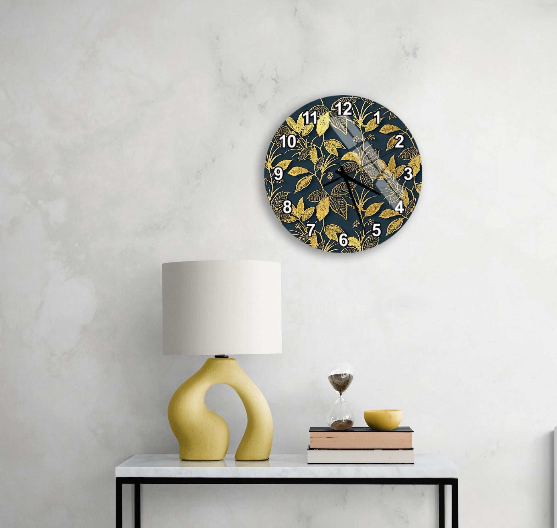 a modern living room with a white marble wall, a round clock with a gold leaf design, a yellow lamp with a unique curved base, and a small table with a book and a glass of wine.