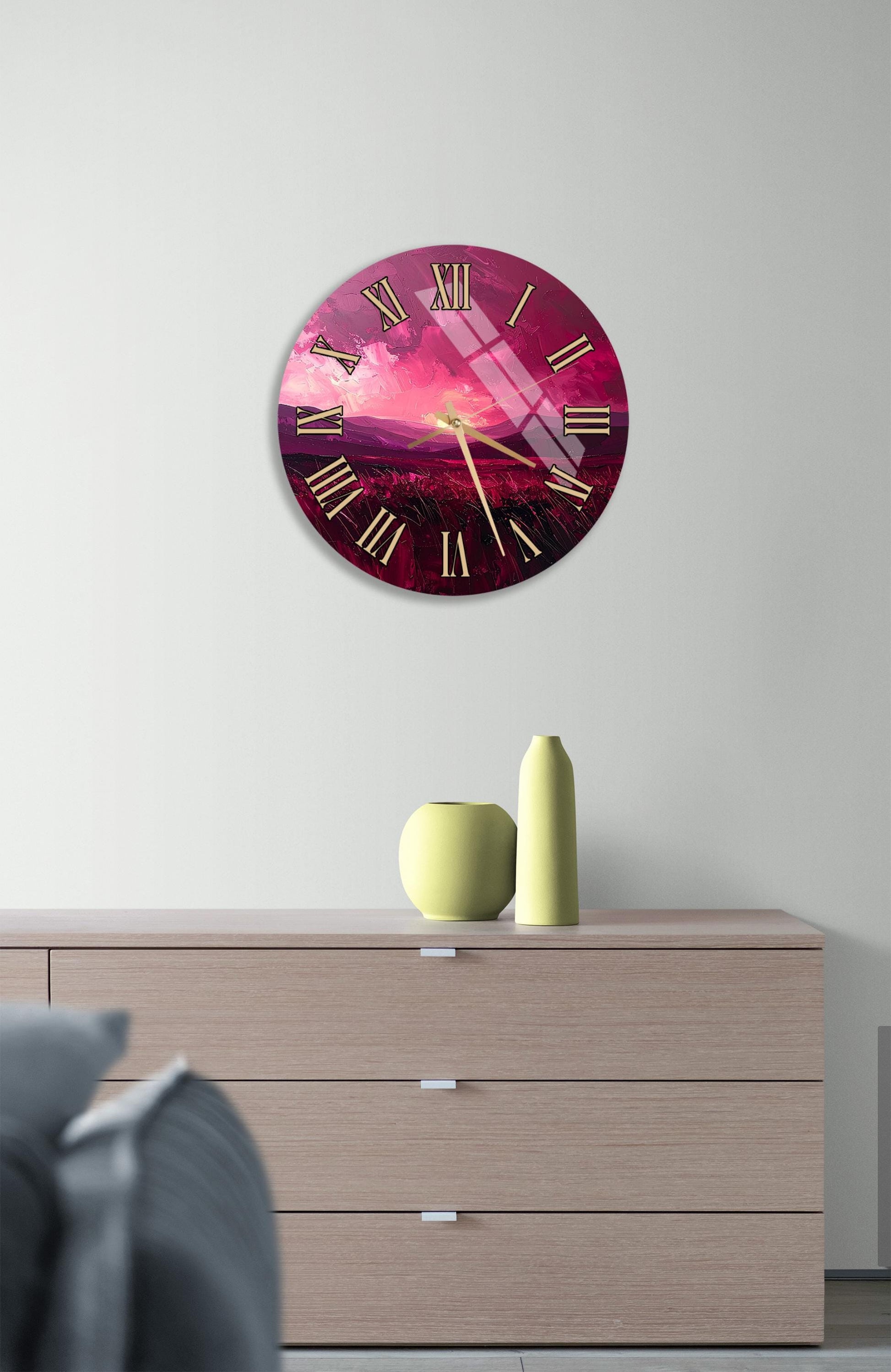a large, round clock with a pink and purple color scheme, mounted on a wall above a wooden dresser. The dresser has three drawers and contains a yellow vase and a small green pot.