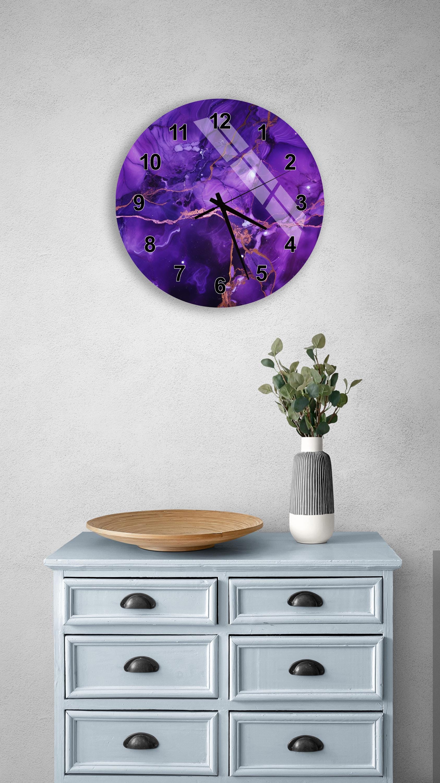 a purple wall clock with a marble-like design, mounted on a wall above a light blue dresser with six drawers. The dresser also has a wooden tray and a vase with greenery on it.