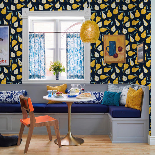a cozy dining area with a white table, blue and white patterned cushions, and a yellow pendant light. A window with blue curtains and a painting of pears is visible, and a vase of flowers is placed on the windowsill. The room has a warm and inviting atmosphere, with a mix of modern and traditional elements.