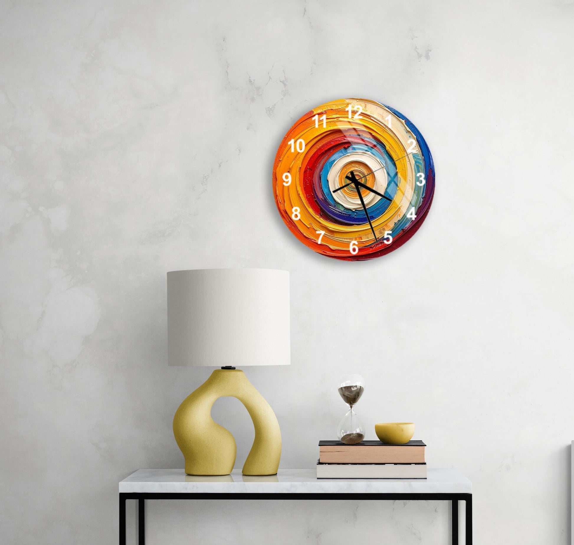 a modern wall clock with a colorful spiral design, mounted on a white wall above a white table with a yellow lamp and a stack of books.