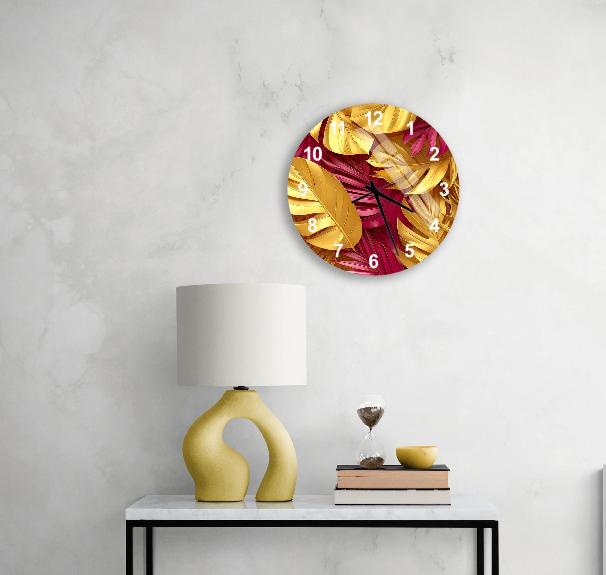 a white wall with a round clock displaying a tropical leaf pattern, a yellow lamp on a white table, and a stack of books and a glass of wine on the table.