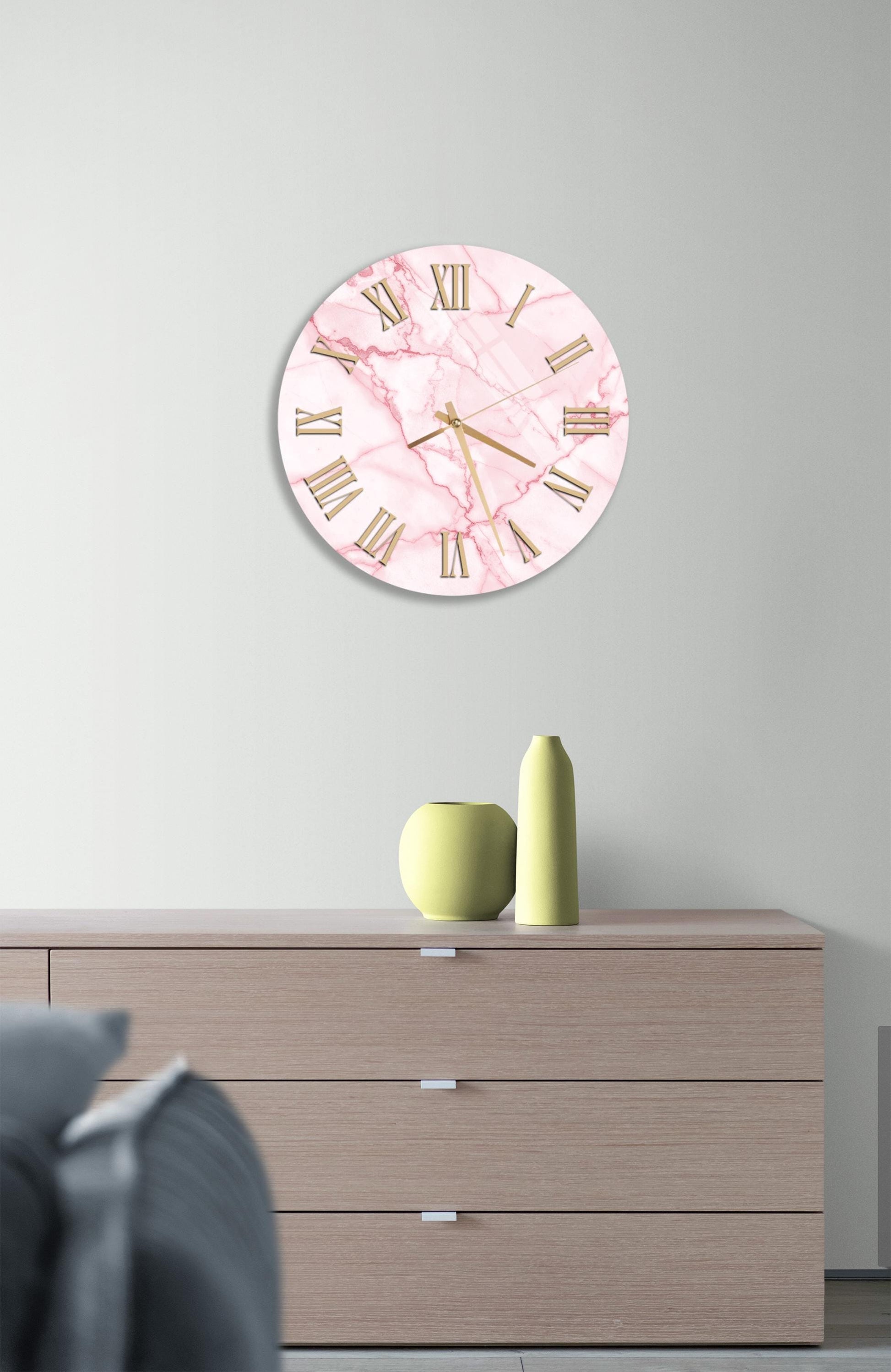 a pink marble clock with Roman numerals on a wall, above a wooden dresser with three drawers.