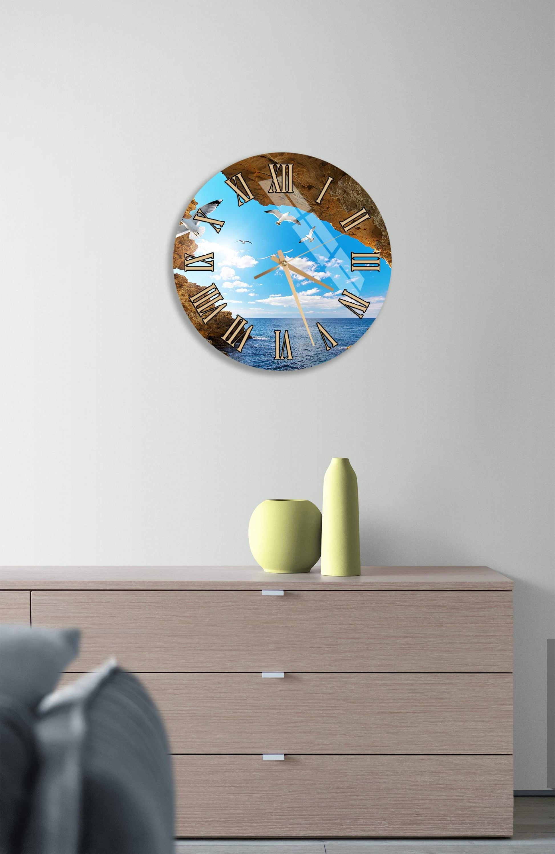 a wall clock with a beach scene on its face, mounted on a wall above a wooden dresser with a vase and a small sculpture on top.