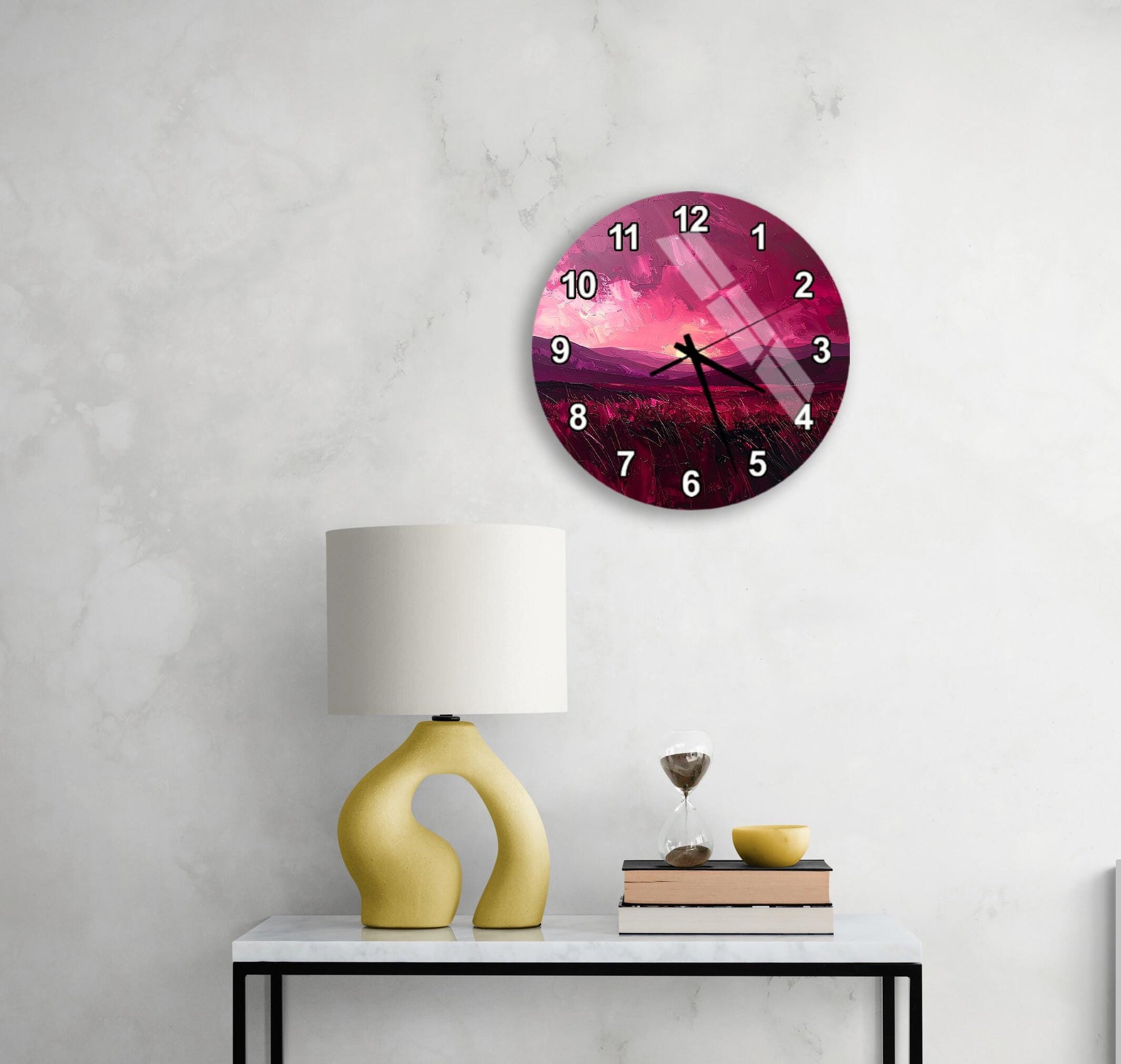 a round wall clock with a pink and purple design, mounted on a wall above a white table with a yellow lamp and a stack of books.