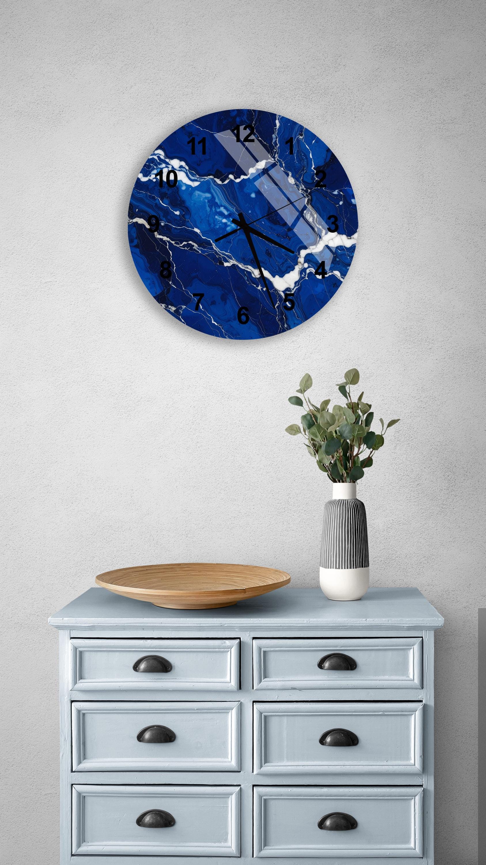 a blue and white marble clock mounted on a wall above a light blue dresser with six drawers. The clock has a marble-like design and displays the time. The dresser also has a vase with greenery and a wooden tray on top.