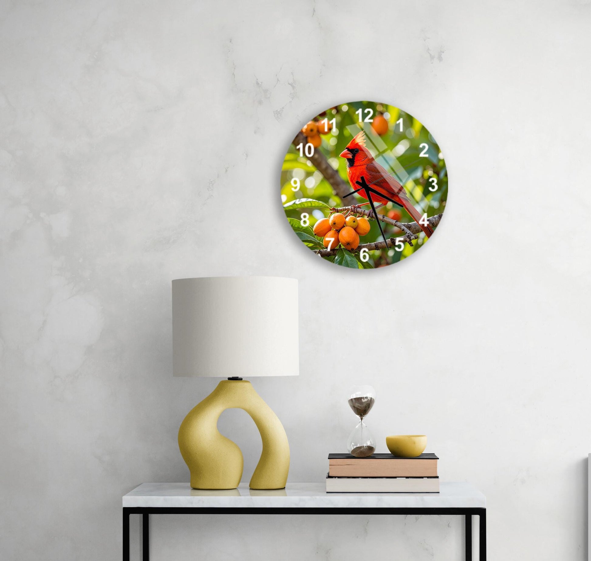 a round clock with a red cardinal perched on a branch, surrounded by orange fruits, placed on a white wall above a white table with a yellow lamp and a glass of water.