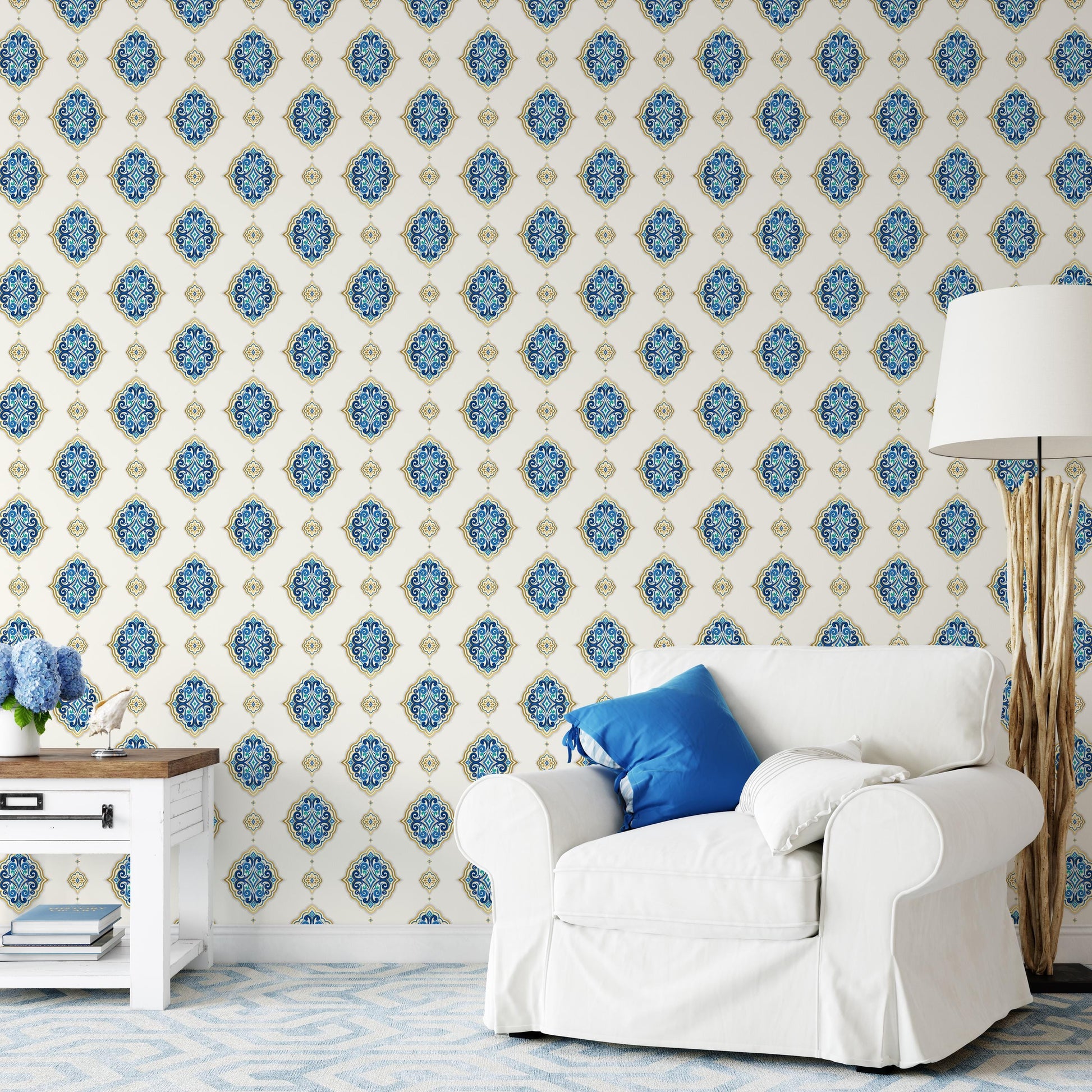 a living room with a white sofa and a wooden side table. The sofa has a blue pillow on it. The wall behind the sofa has a repeating pattern of blue and gold floral designs. There is also a lamp on the side table.