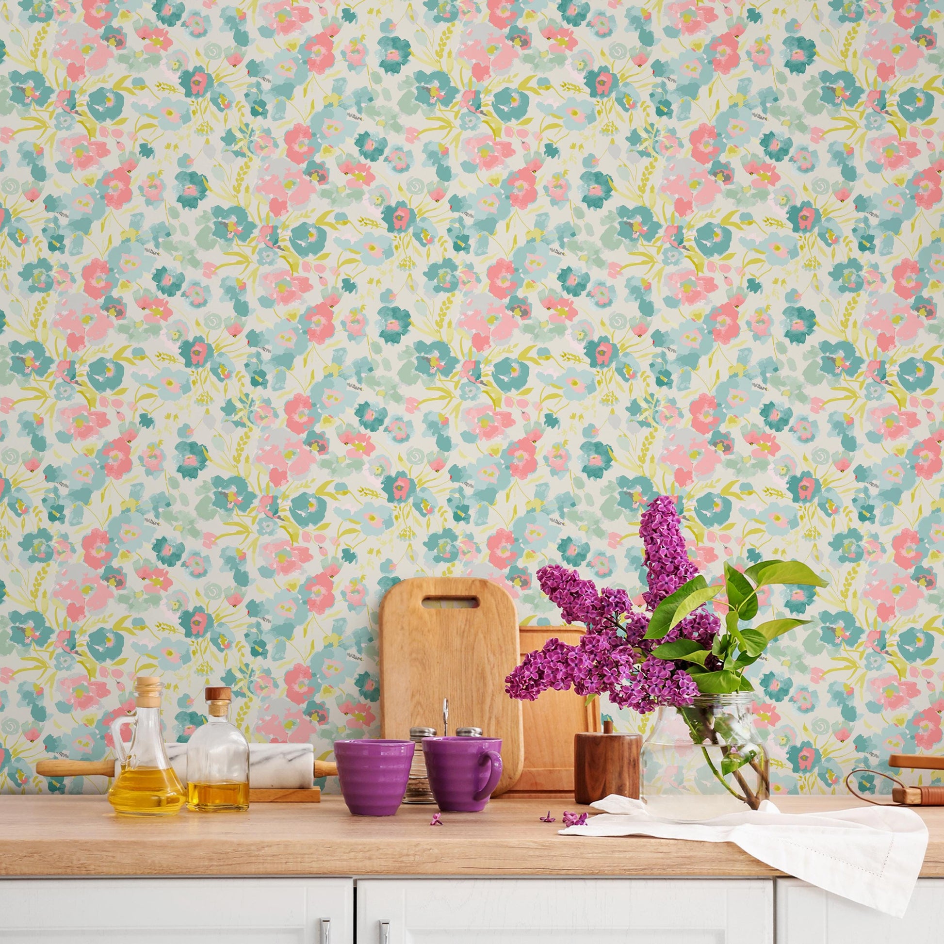 a kitchen counter with a floral wallpaper, a vase of purple flowers, and various kitchen items such as a wooden cutting board, a glass bottle, and a cup.