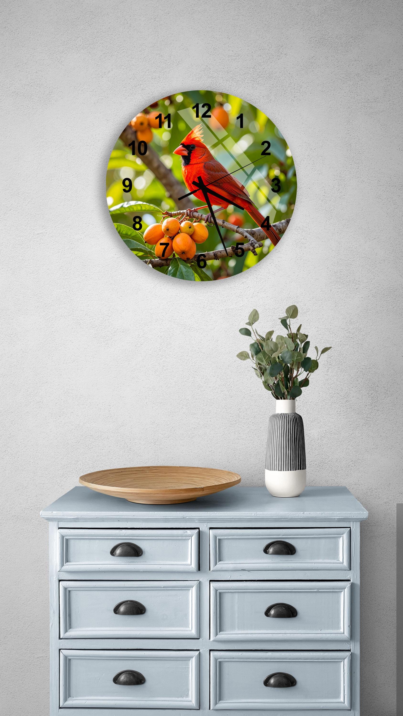 a round clock with a red cardinal perched on a branch, surrounded by orange berries, hanging on a wall above a light blue dresser with six drawers.