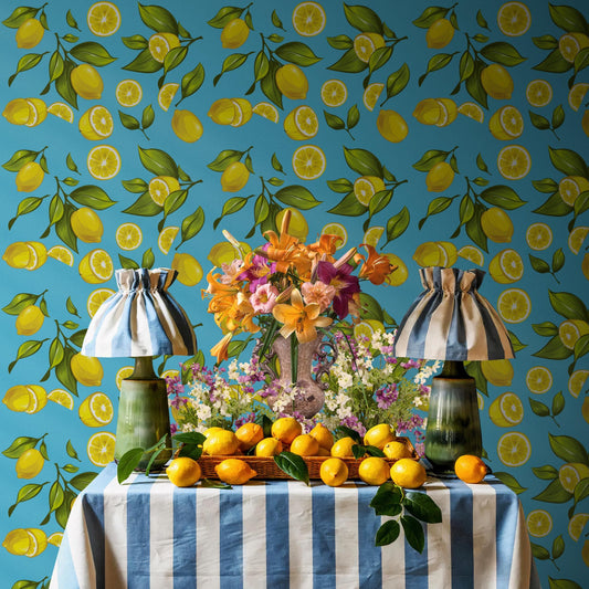 a table with a blue and white striped tablecloth, a vase of flowers, and a basket of oranges. The table is set against a wall with a pattern of yellow and green lemons.