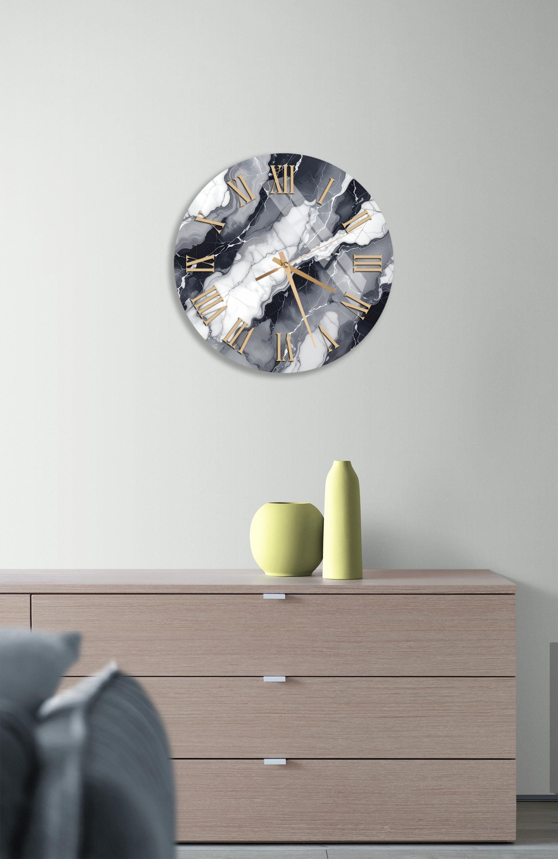 a modern interior design with a round clock featuring a marble-like pattern on the wall above a wooden dresser with three drawers. The dresser also has a yellow vase and a green vase on it, adding a pop of color to the neutral-toned room.