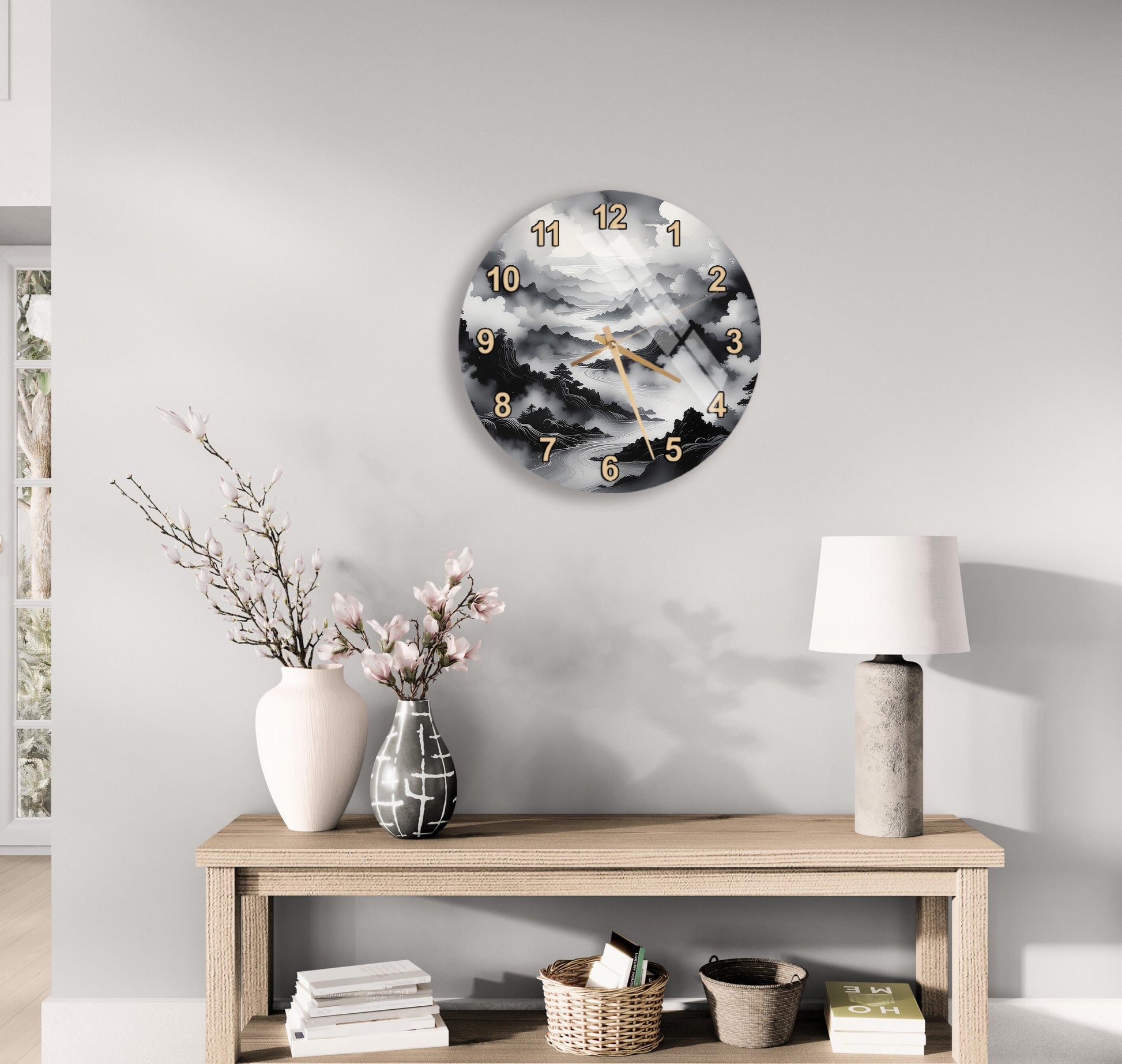 a wall clock with a black and white landscape design, a wooden table with a lamp, a vase with pink flowers, and a stack of books.