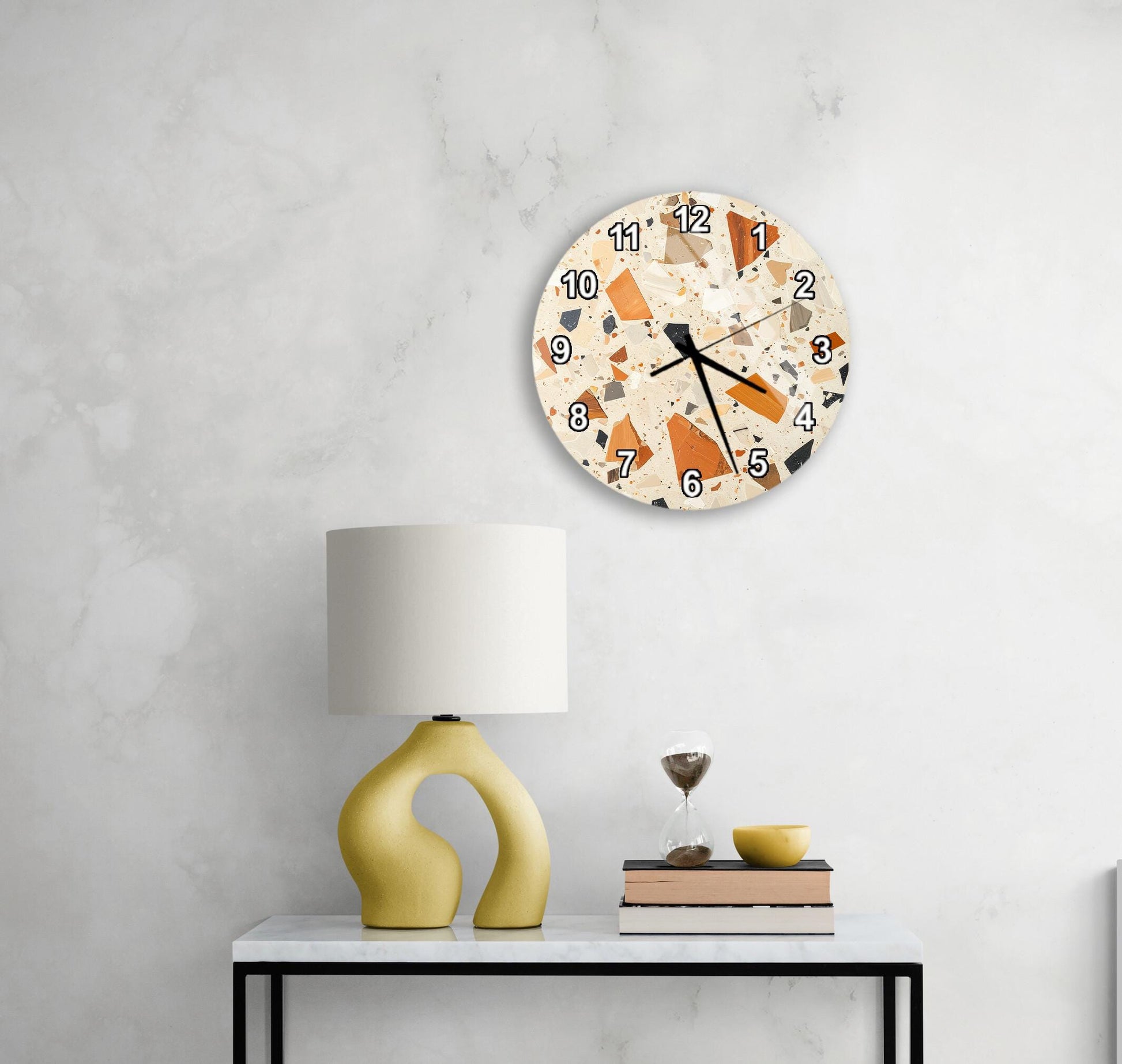 a modern, abstract-style clock with a unique design hanging on a wall above a white table with a yellow lamp and a stack of books.