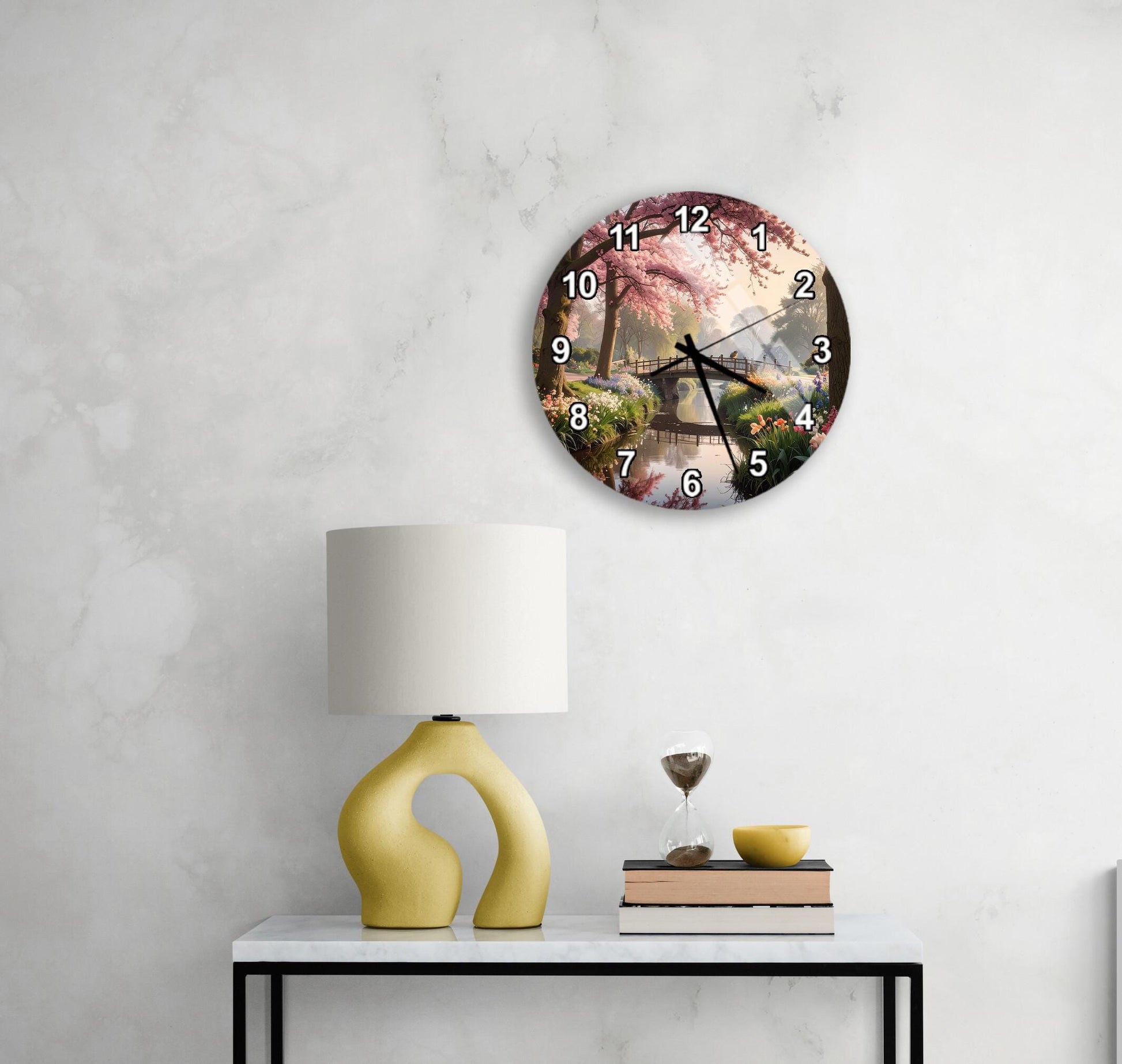 a white wall with a round clock displaying a scenic image of a garden with pink flowers and a tree. Below the clock, there is a yellow lamp on a white table, and a stack of books and a glass of wine are also present on the table.