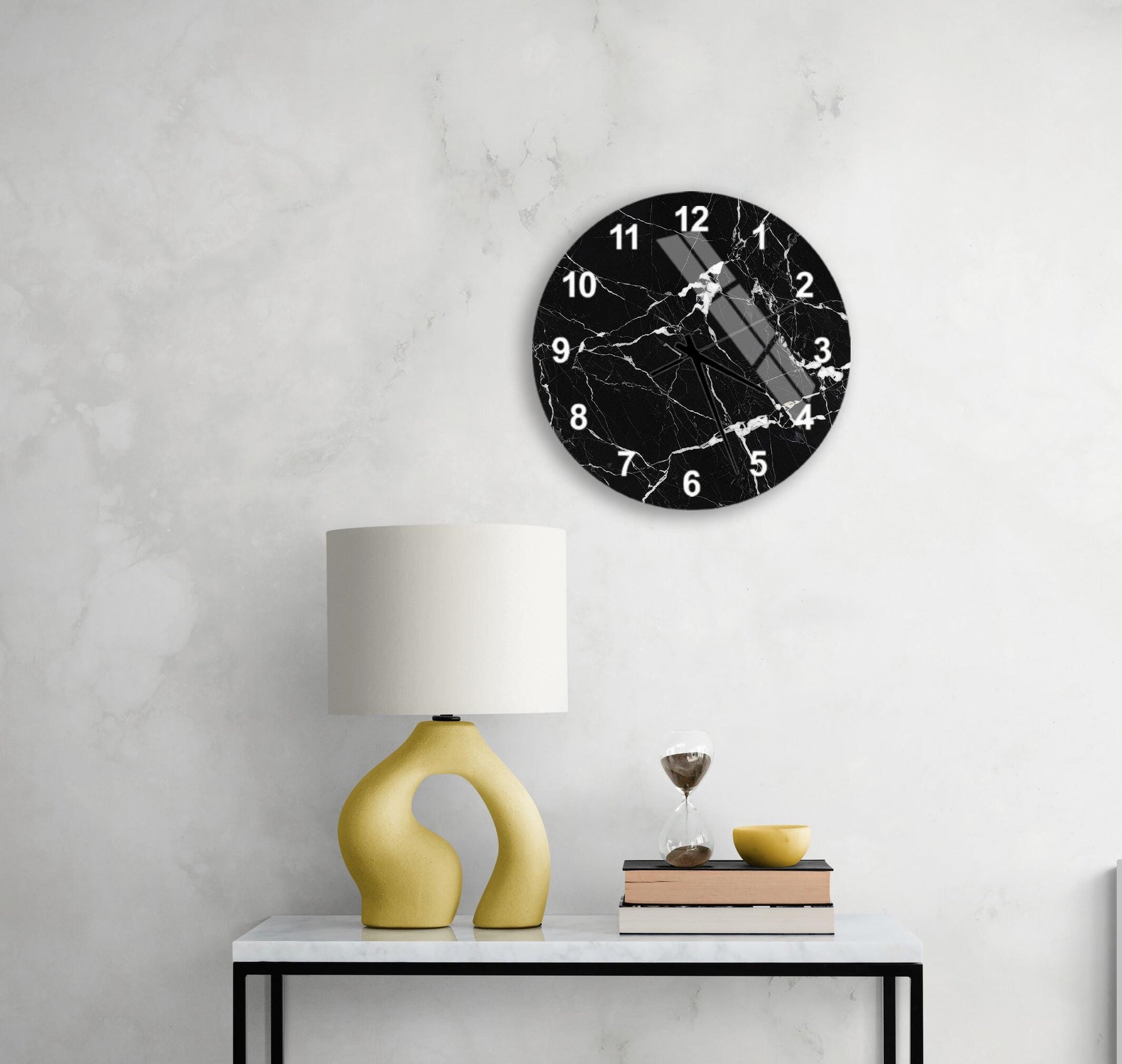 a black and white marble clock mounted on a white wall above a white table with a yellow lamp and a stack of books.