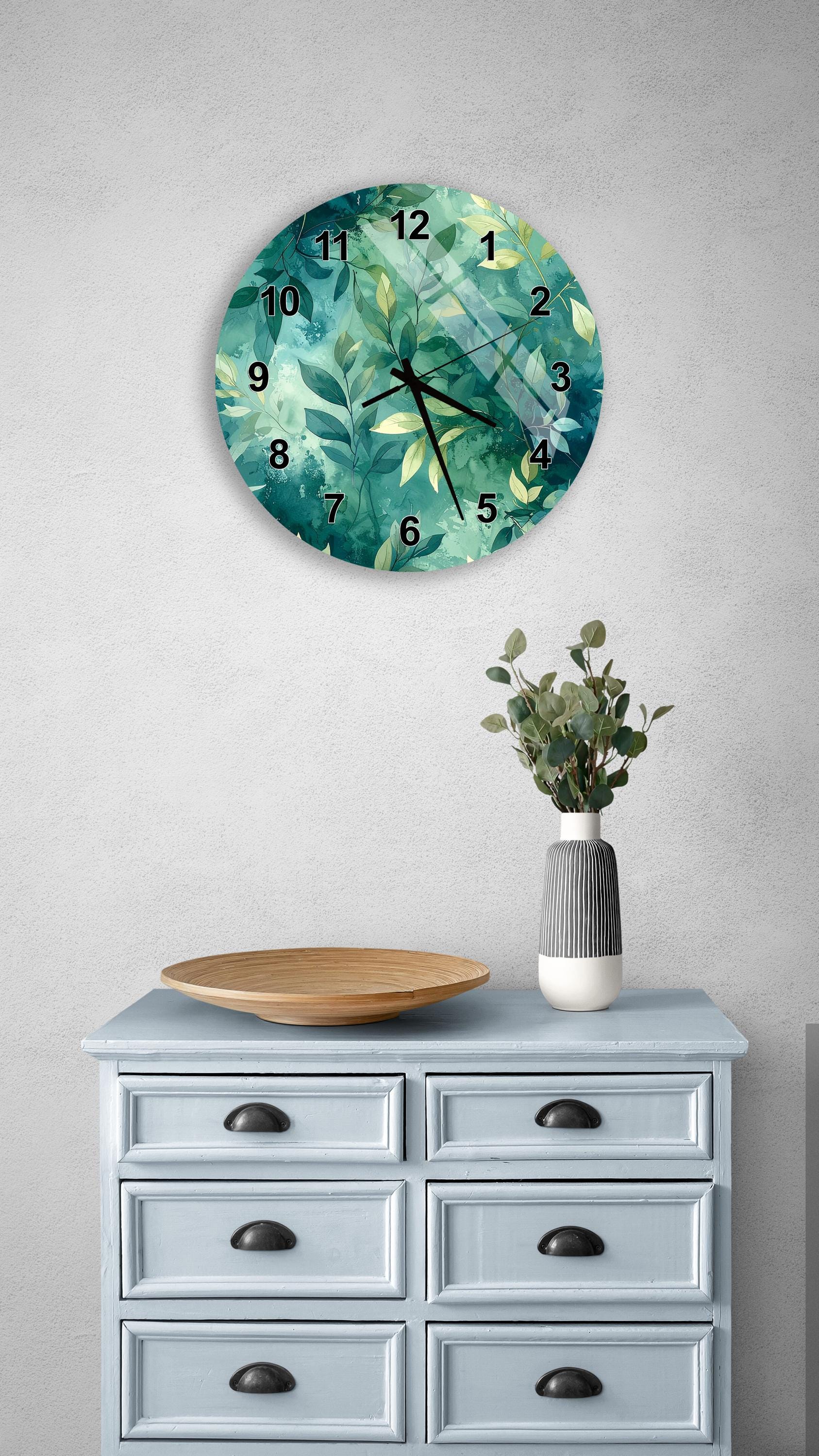 a wall clock with a green leaf design, a wooden bowl on a light blue dresser, and a vase with greenery on a gray wall.