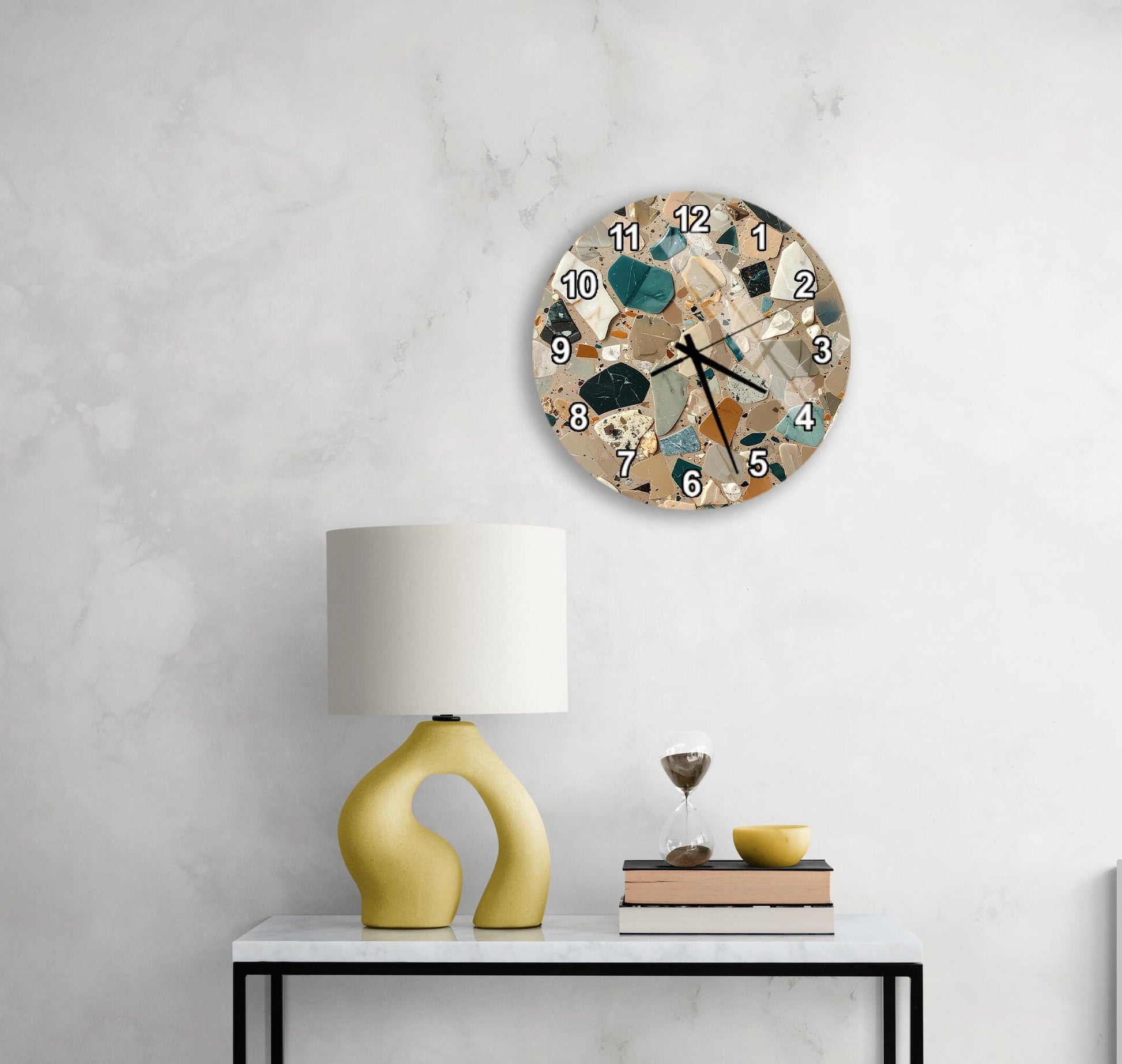 a modern, abstract-style clock with a mosaic design on the wall above a white table with a yellow lamp and a stack of books.