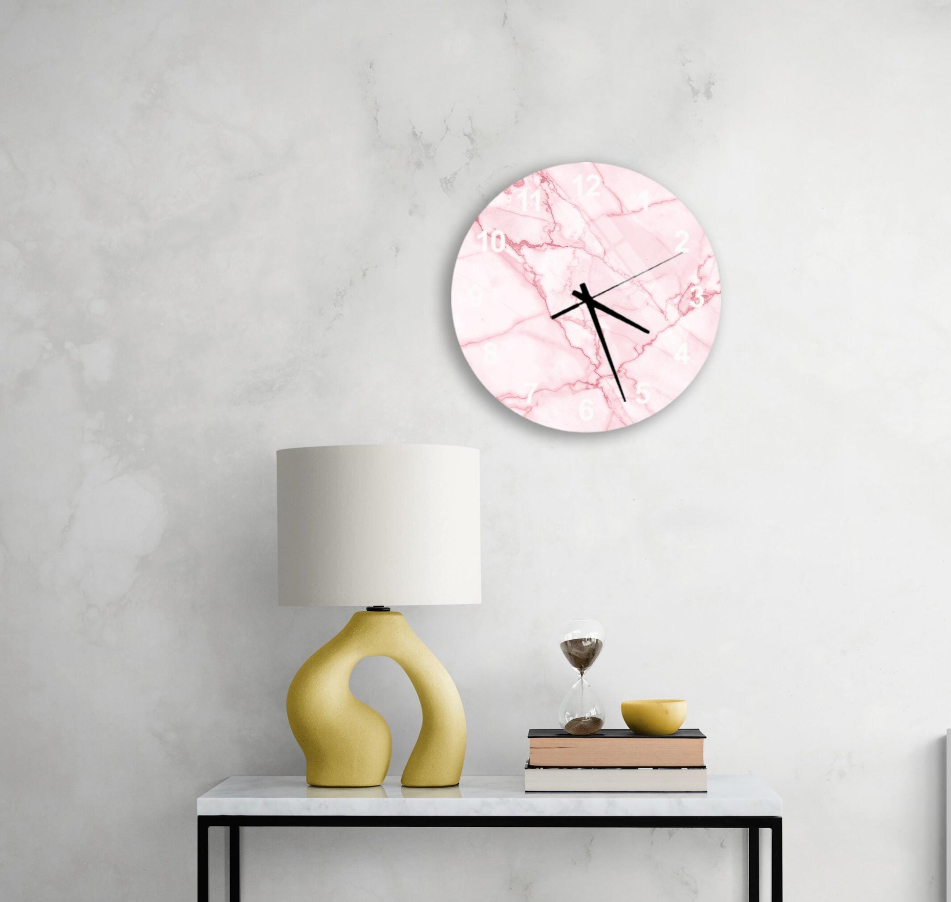 a white marble wall with a pink marble clock, a yellow lamp, a stack of books, and a glass of wine on a white table.