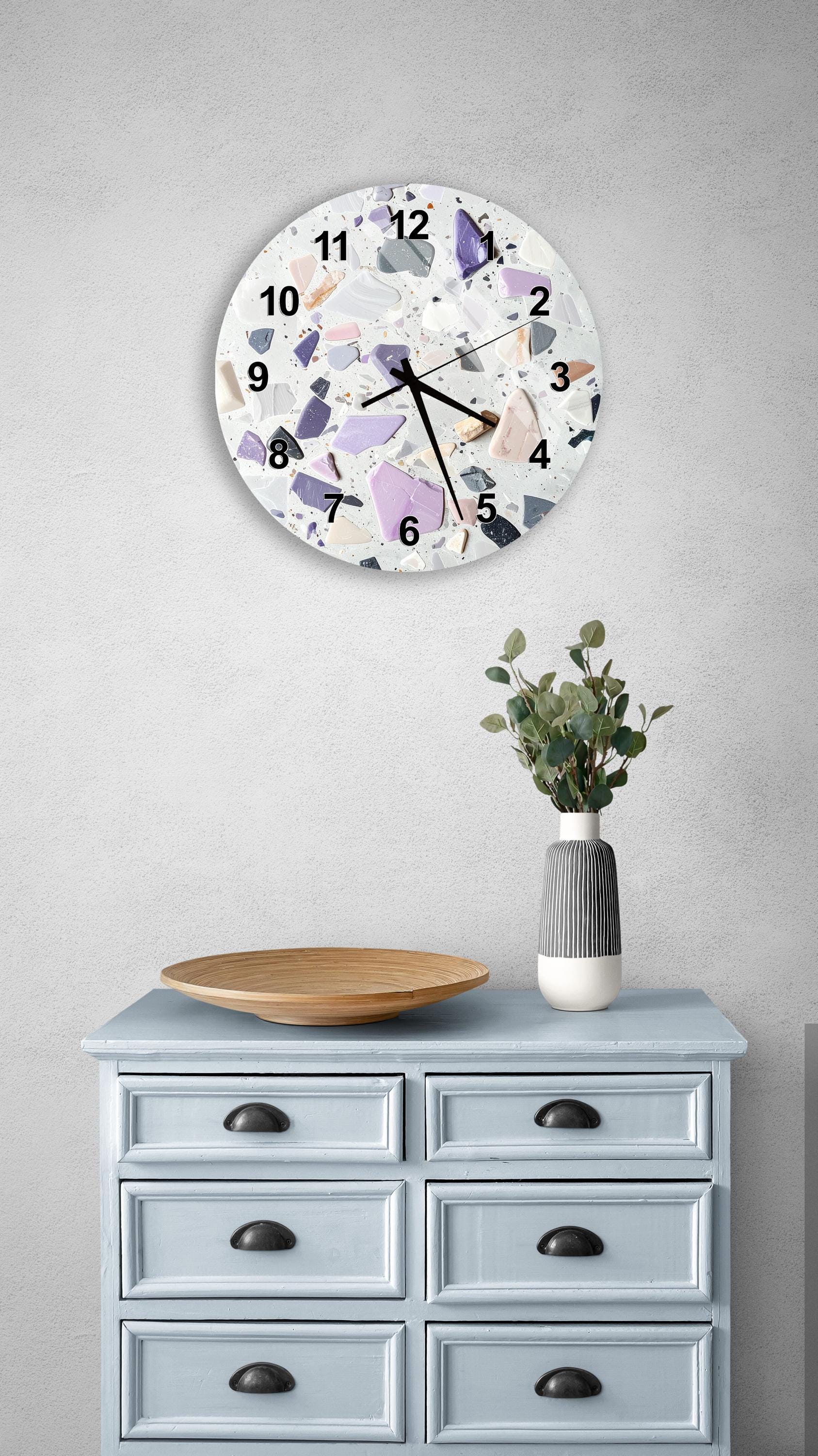 a wall clock with a unique, colorful design, mounted on a wall above a light blue dresser with six drawers. The dresser also has a vase with greenery on it, and there is a wooden bowl on the dresser.