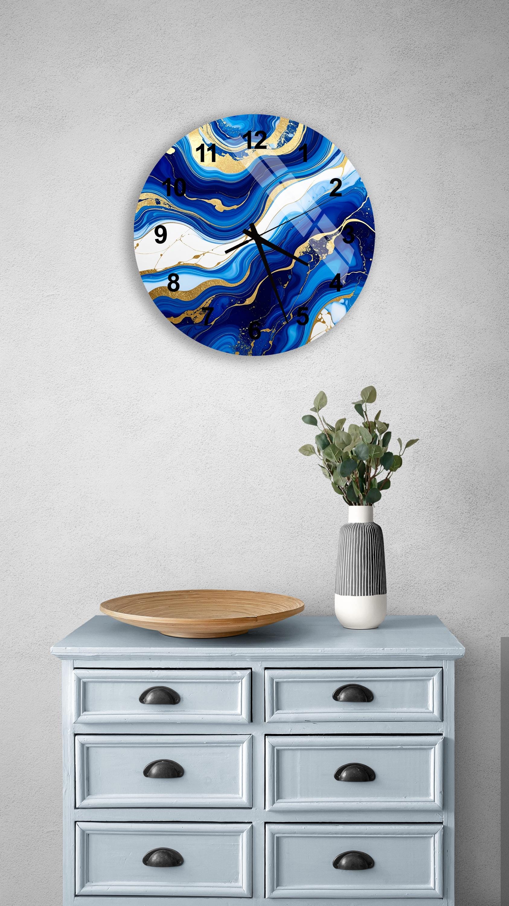 a wall clock with a blue and white marble design, mounted on a wall above a light blue dresser with six drawers. The dresser also has a vase with greenery on it, and there is a wooden bowl on top of the dresser.