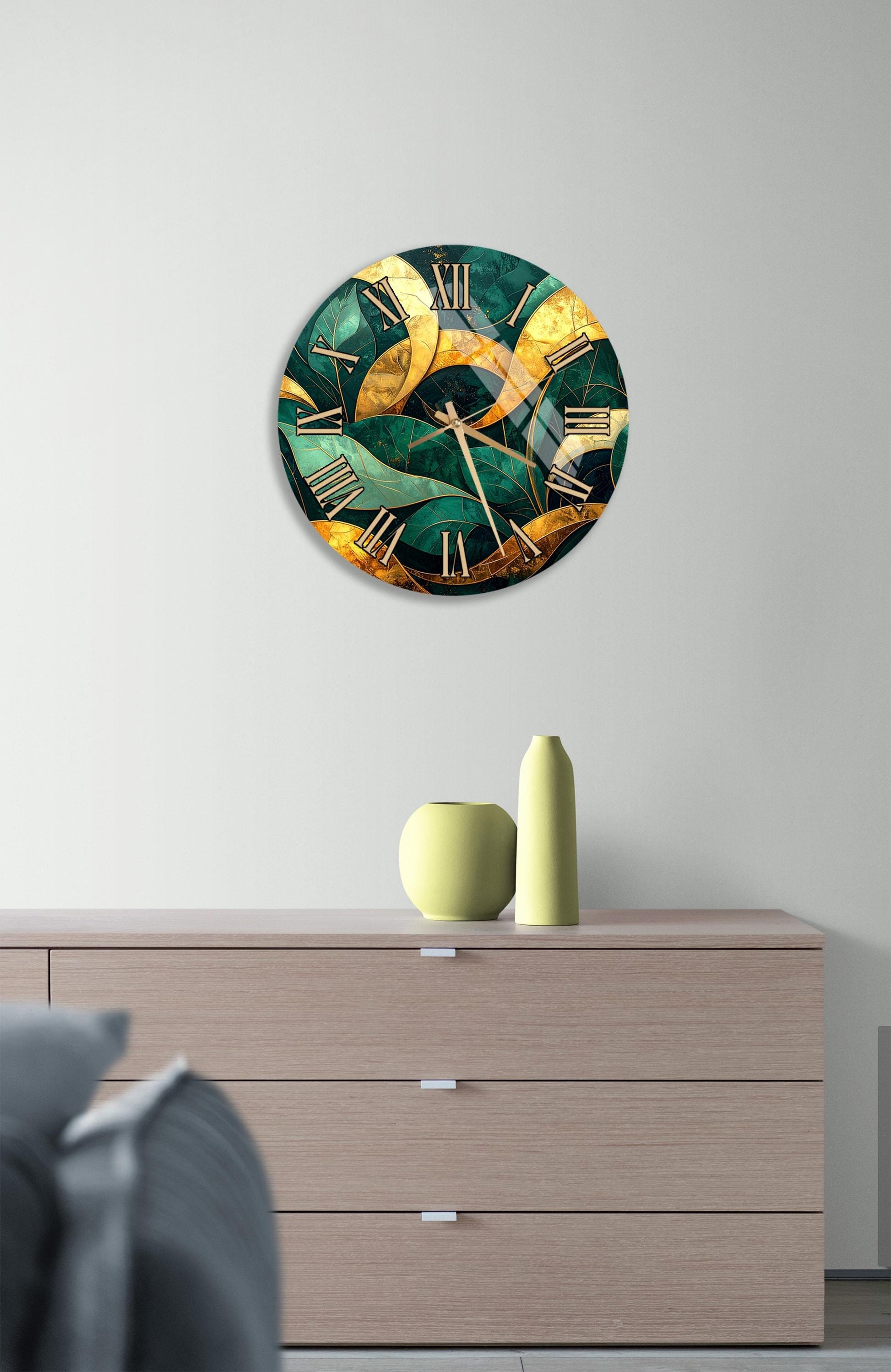 a modern clock with a green and gold design, mounted on a wall above a wooden dresser with a vase and a small sculpture on top.