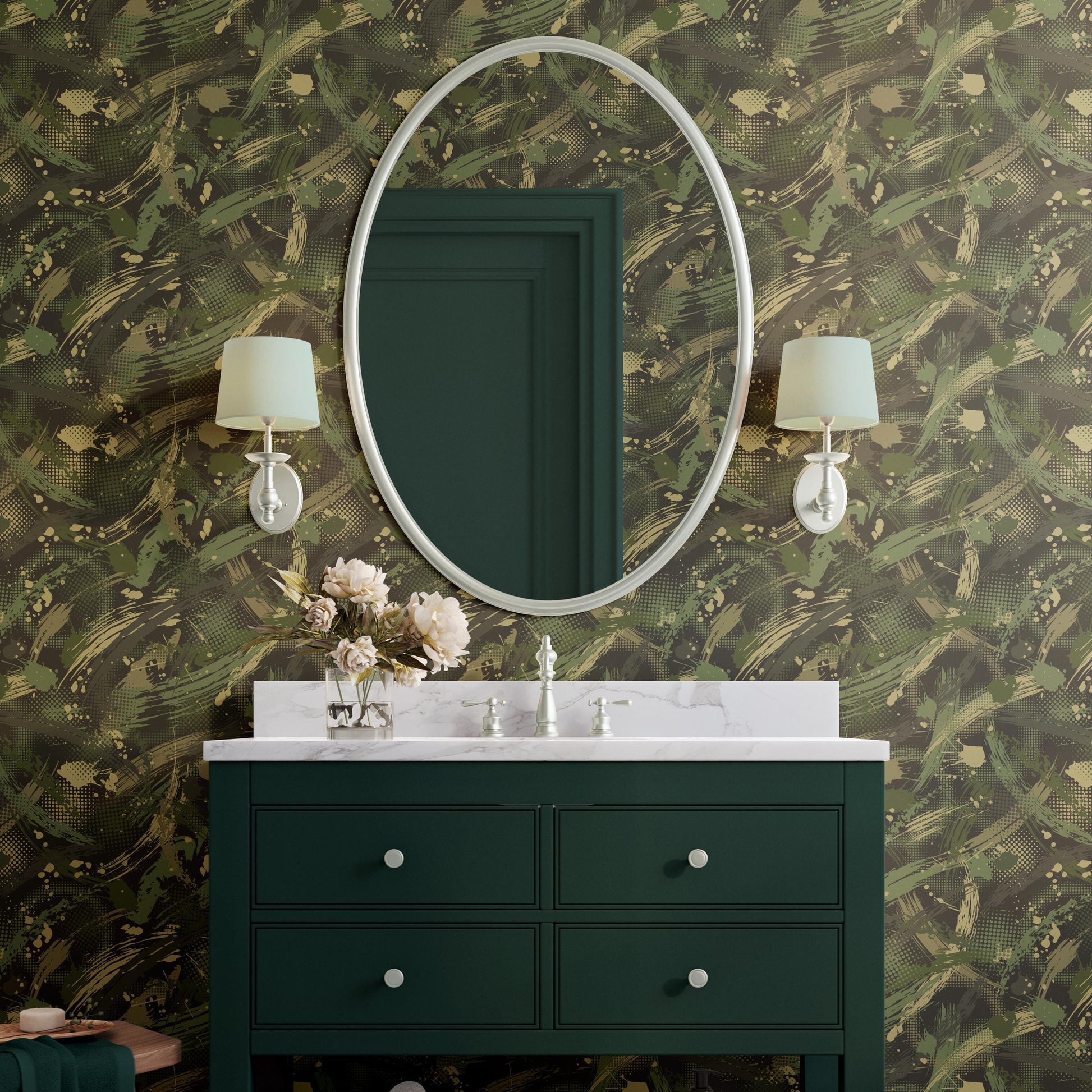 a bathroom vanity with a green cabinet, a white countertop, and a large oval mirror above it. The wall behind the vanity has a camouflage pattern. There are two wall sconces on either side of the mirror, and a vase of flowers is placed on the countertop.