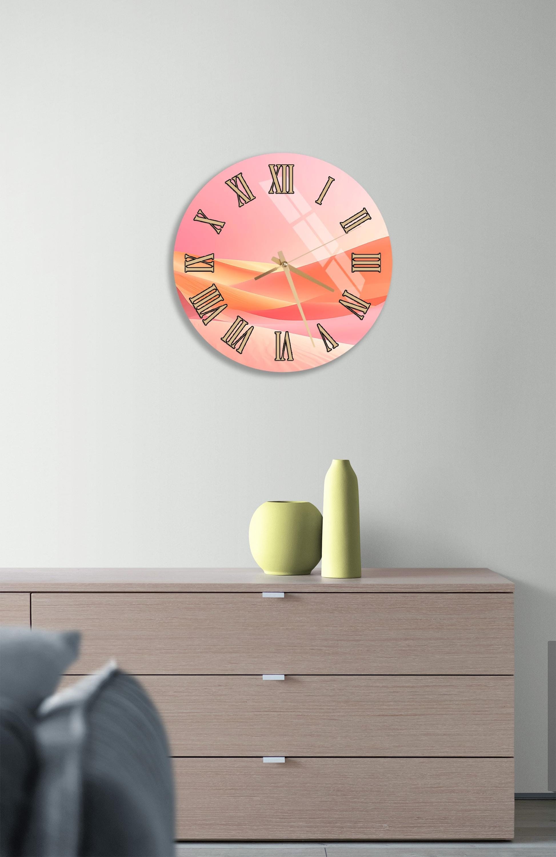 a pink wall clock with Roman numerals, mounted on a wall above a wooden dresser with three drawers.