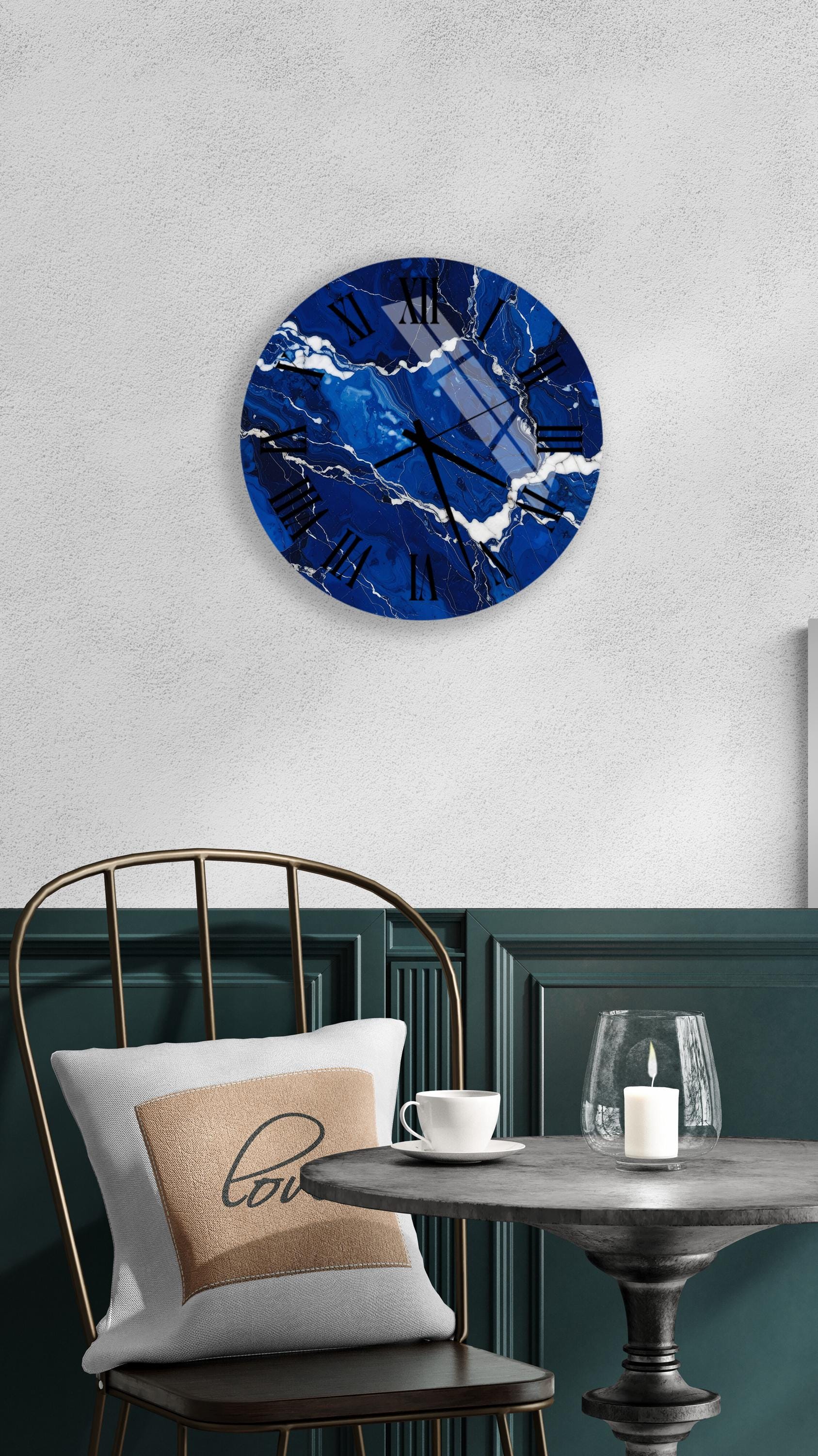 a modern interior design with a blue marble clock as the focal point. The clock is mounted on a white wall above a black chair with a gold frame. The chair is positioned in front of a round table with a white tablecloth. On the table, there is a white coffee cup and a lit candle, creating a cozy and inviting atmosphere.