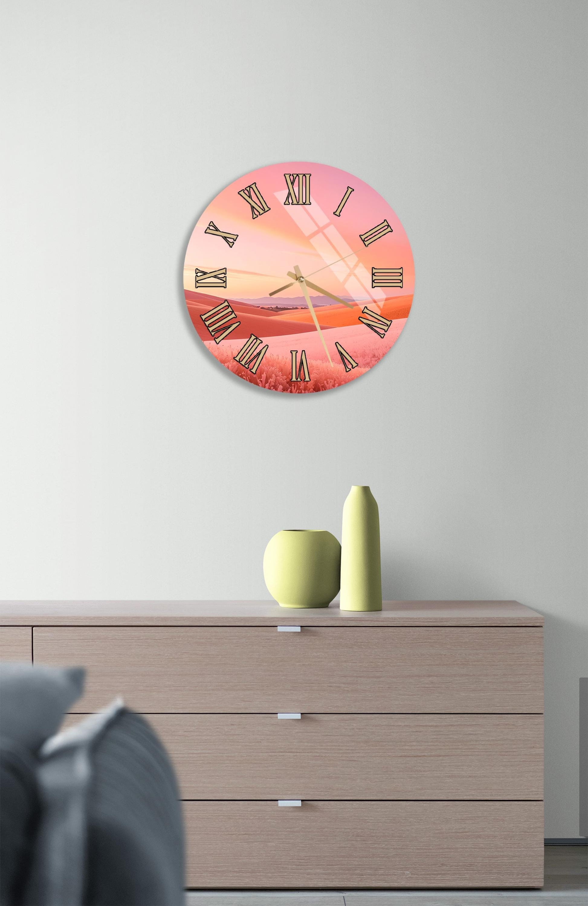 a wall clock with a pink and orange sunset design, mounted on a wall above a wooden dresser with a vase and a small sculpture on top.