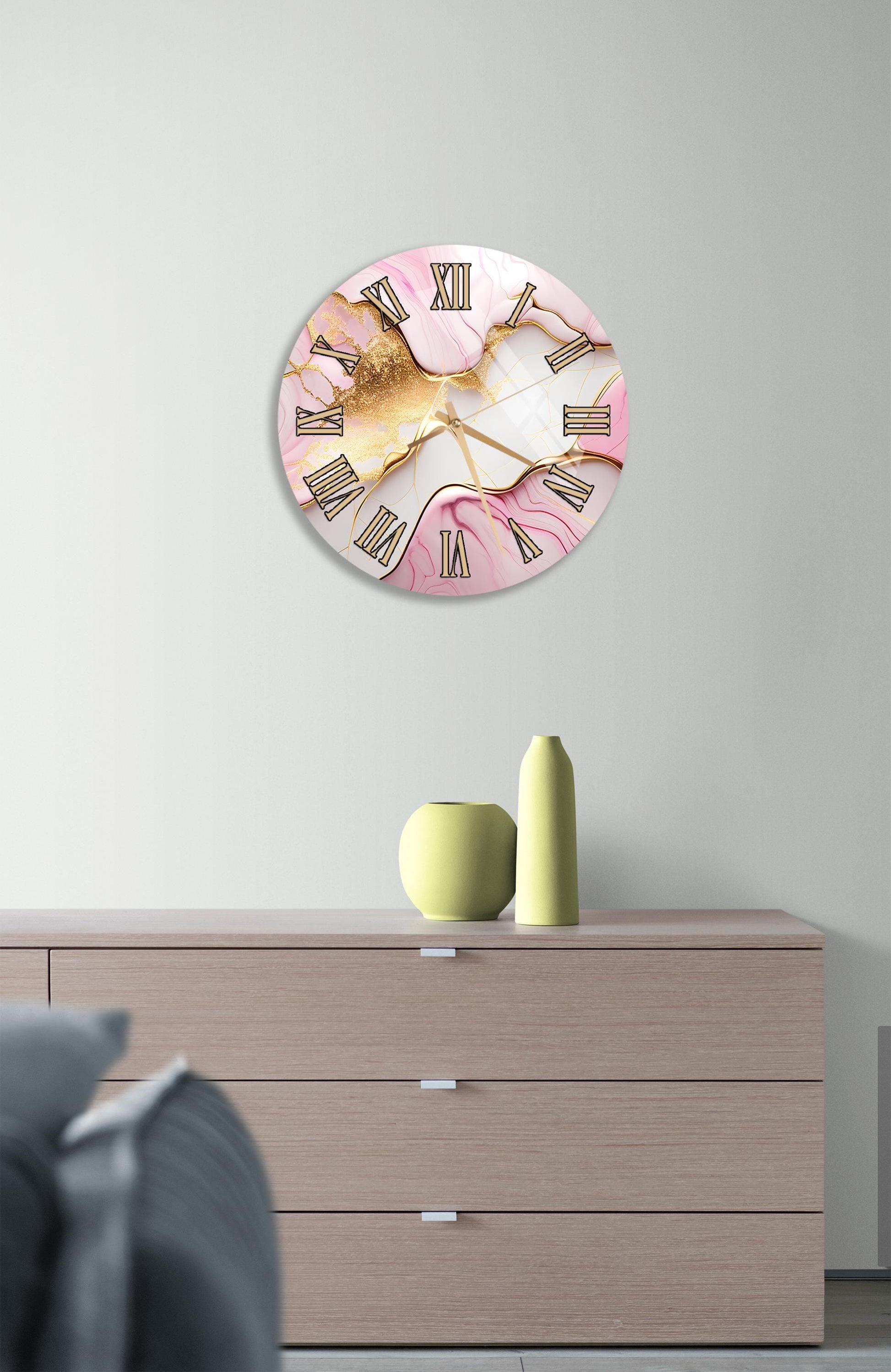 a wall clock with a pink and gold design, mounted on a wall above a wooden dresser with a vase and a small green pot on top.