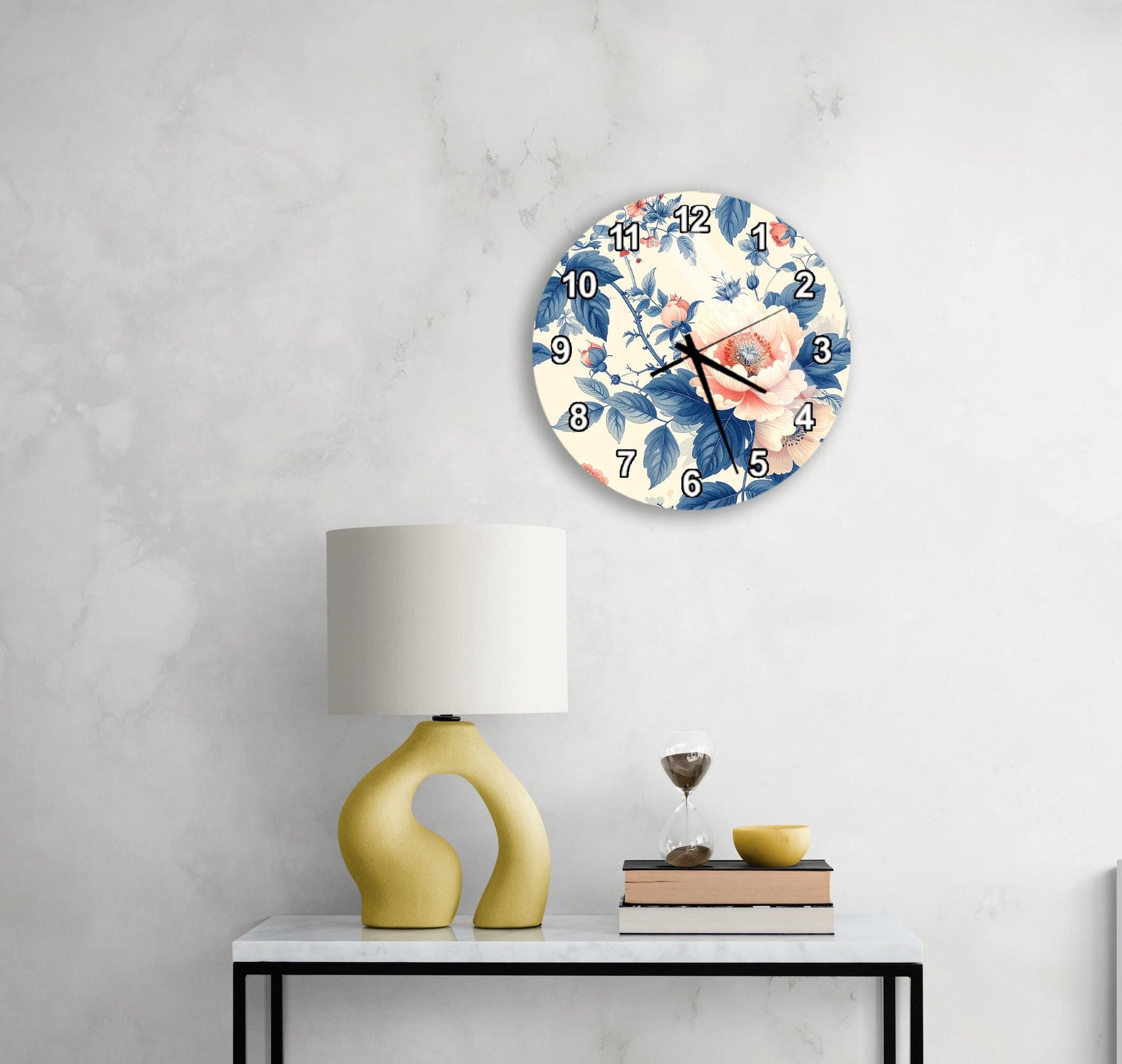 a white wall with a round clock displaying a floral design, a yellow lamp on a white table, and a stack of books and a glass of wine on the table.