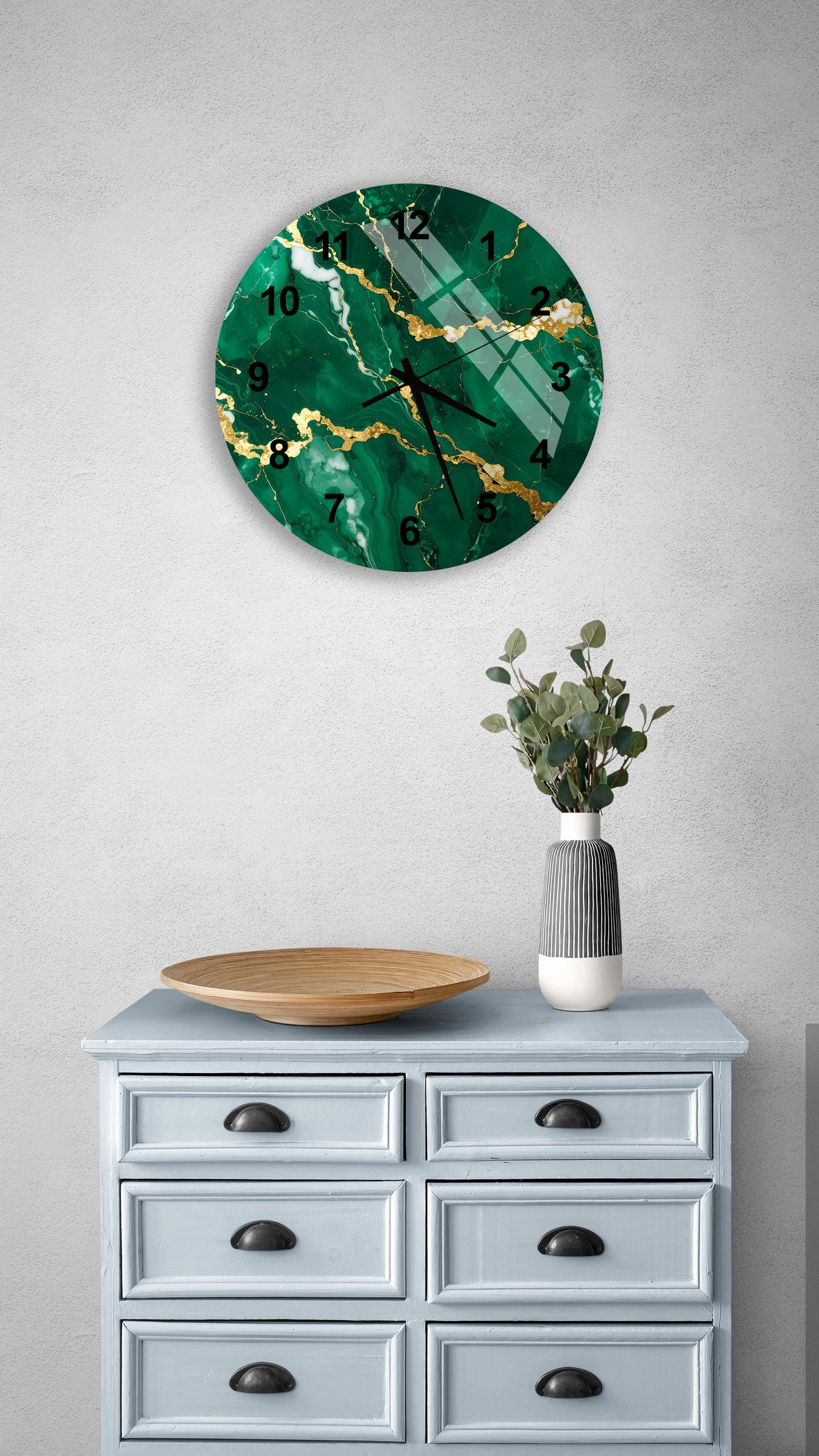 a wall clock with a green marble-like design, mounted on a wall above a light blue dresser with six drawers. The dresser also has a vase with greenery and a wooden bowl on it.