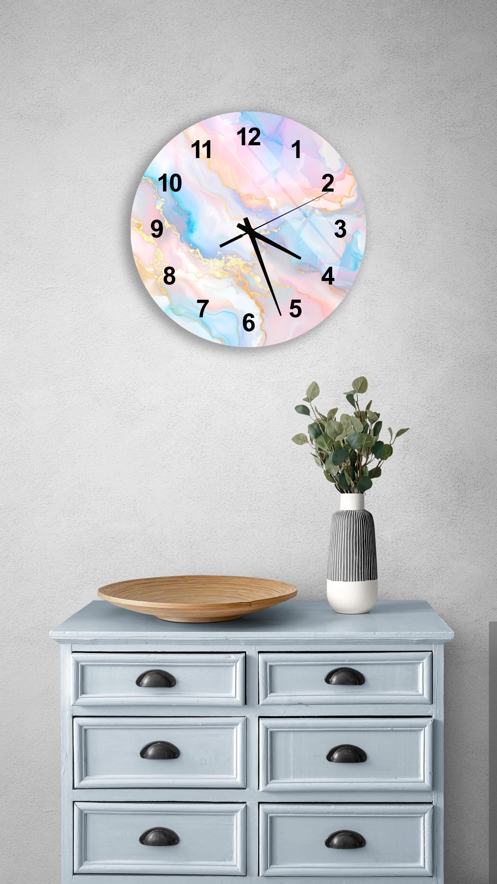 a wall clock with a colorful, abstract design, mounted on a wall above a light blue dresser with six drawers. The dresser also has a vase with greenery on it, and there is a wooden bowl on the dresser.