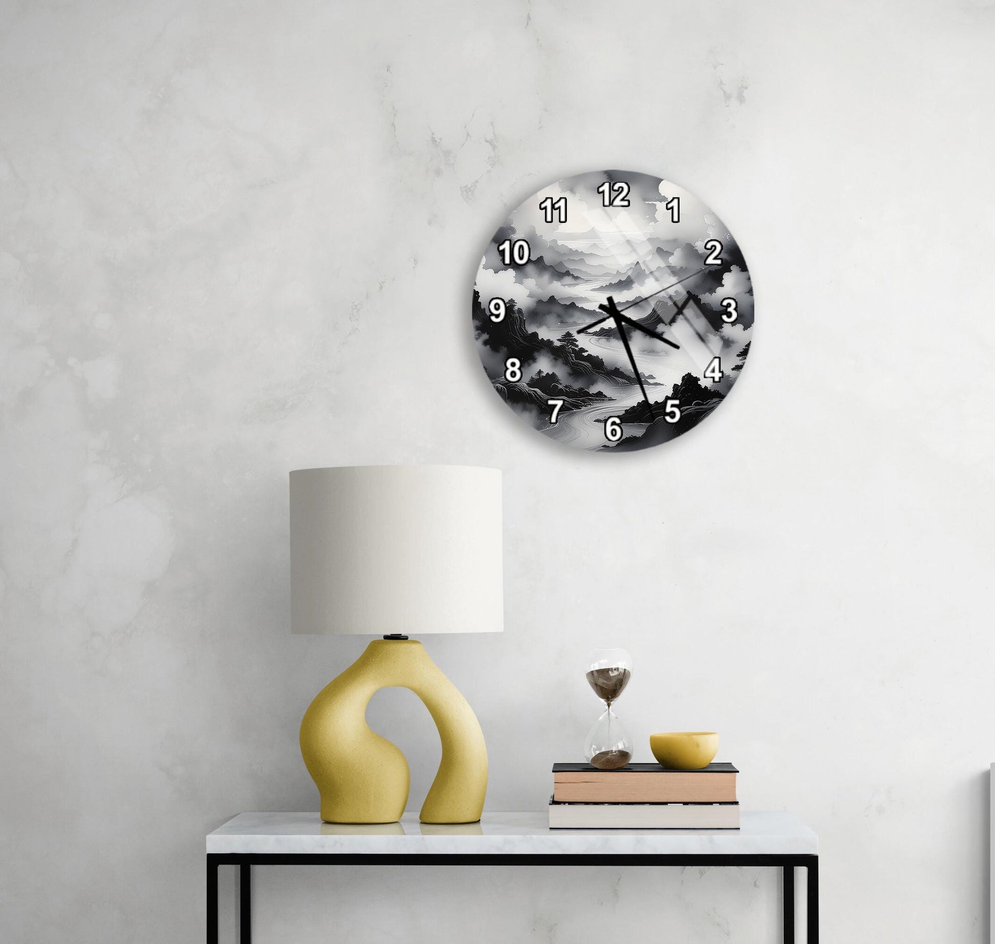 a modern black and white clock with a landscape scene on its face, mounted on a wall above a white table with a yellow lamp and a stack of books.