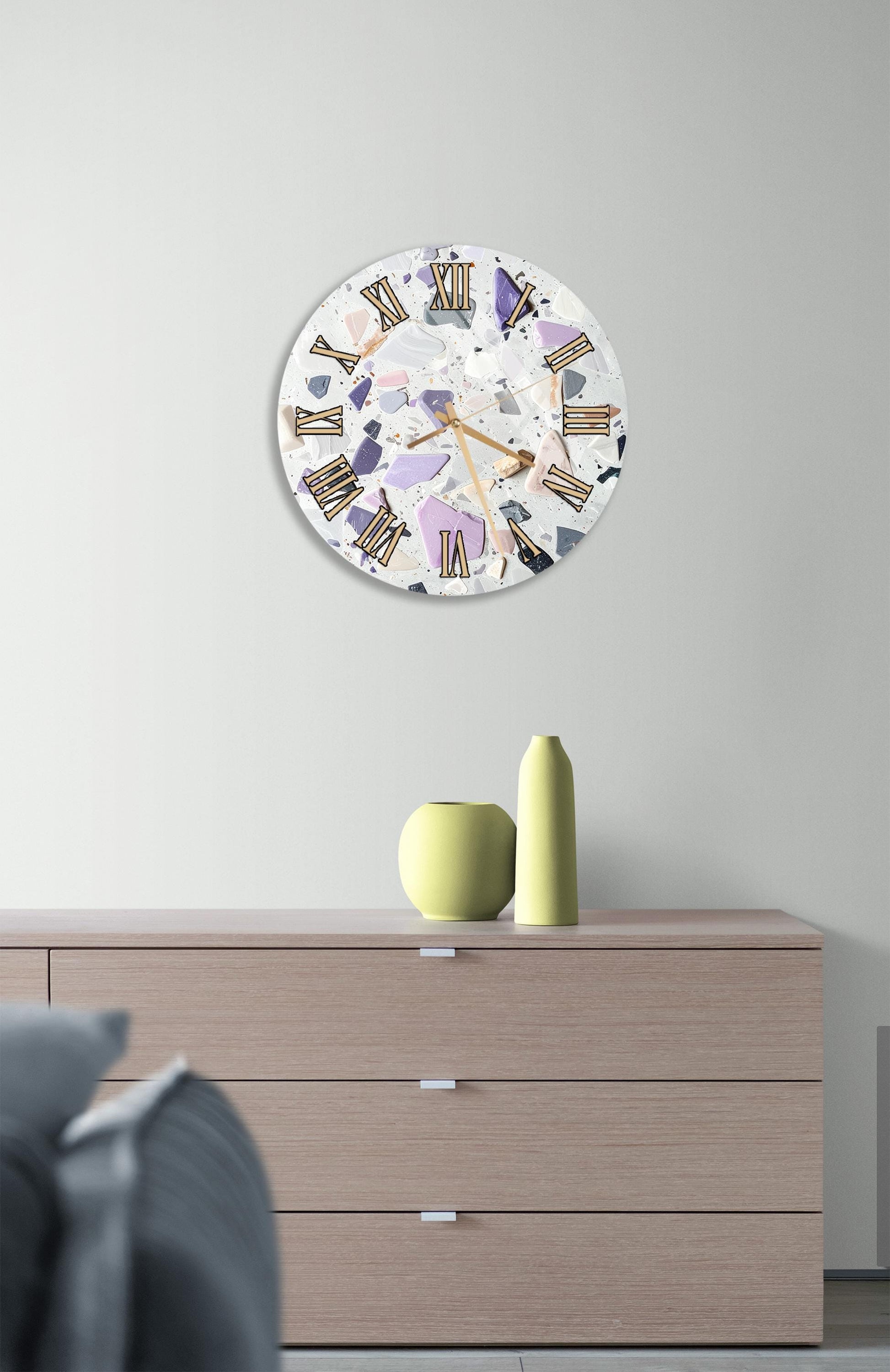 a modern clock with a white face and colorful Roman numerals, mounted on a wall above a wooden dresser.