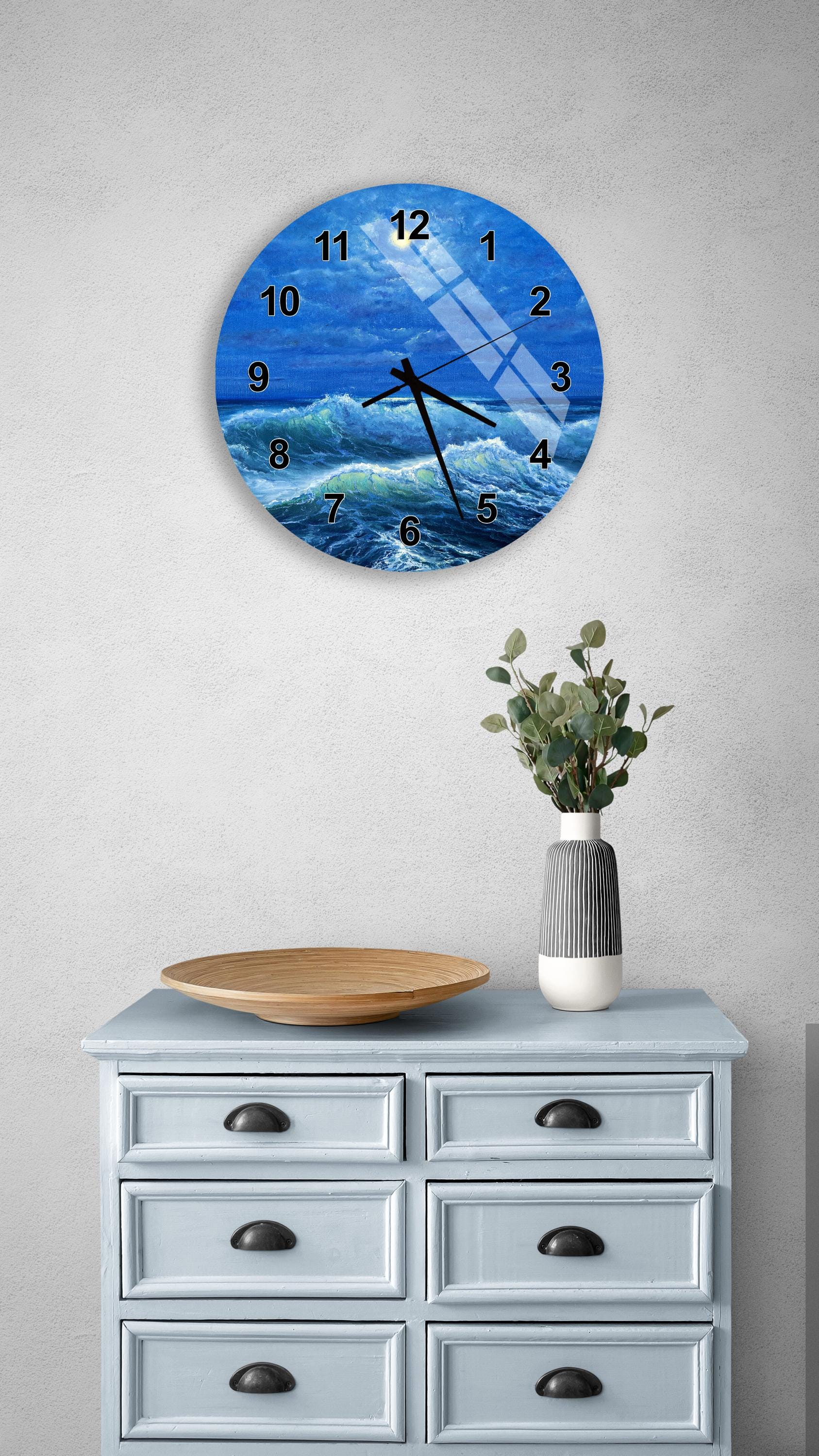a round wall clock with a blue ocean scene on its face, mounted on a wall above a light blue dresser with a wooden tray and a vase of greenery.