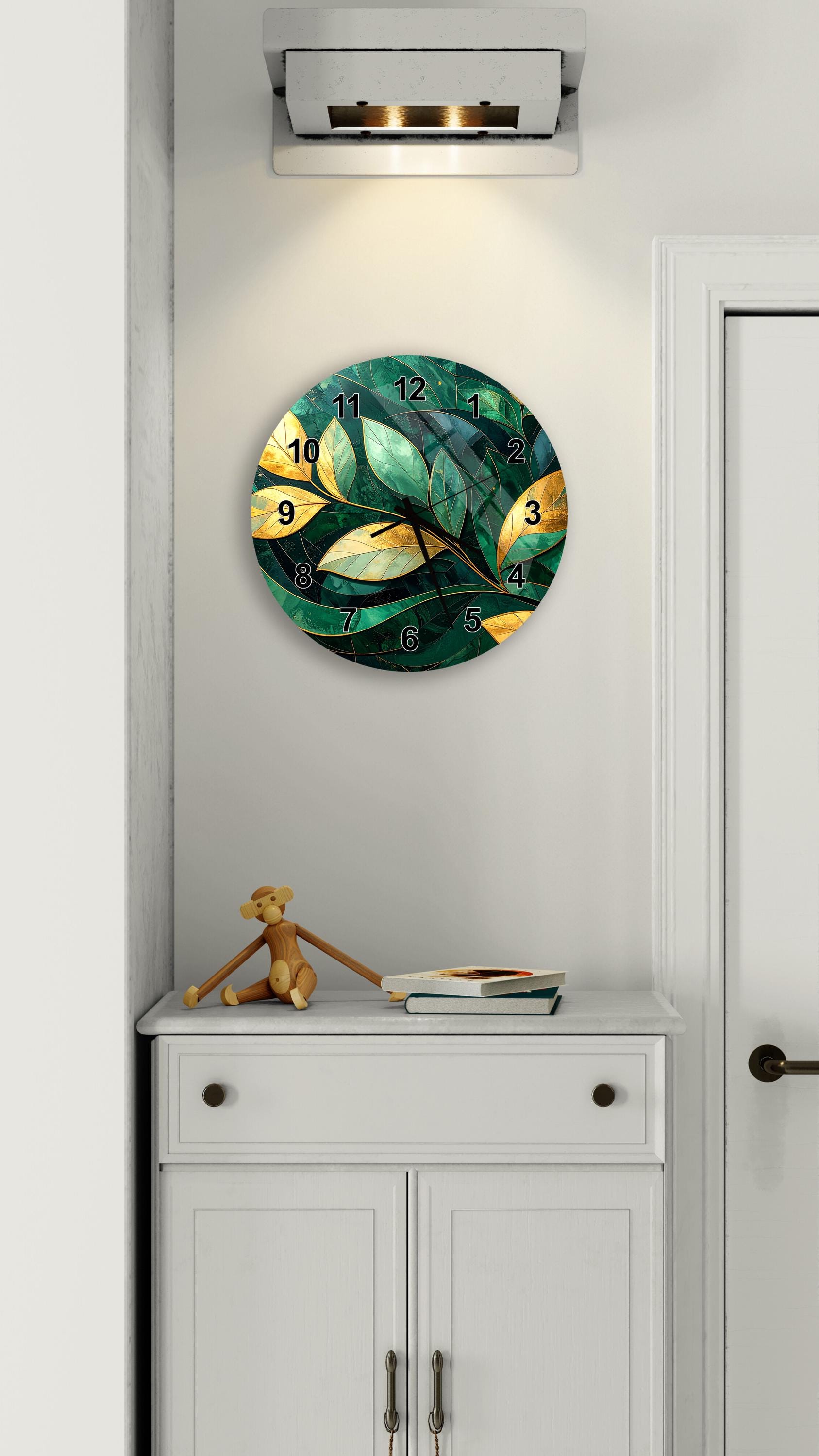 a wall clock with a green leaf design, mounted on a white wall above a white cabinet. The clock has a gold leaf design and displays the numbers 1 through 12. A small brown teddy bear is sitting on top of the cabinet, and there is a book nearby.