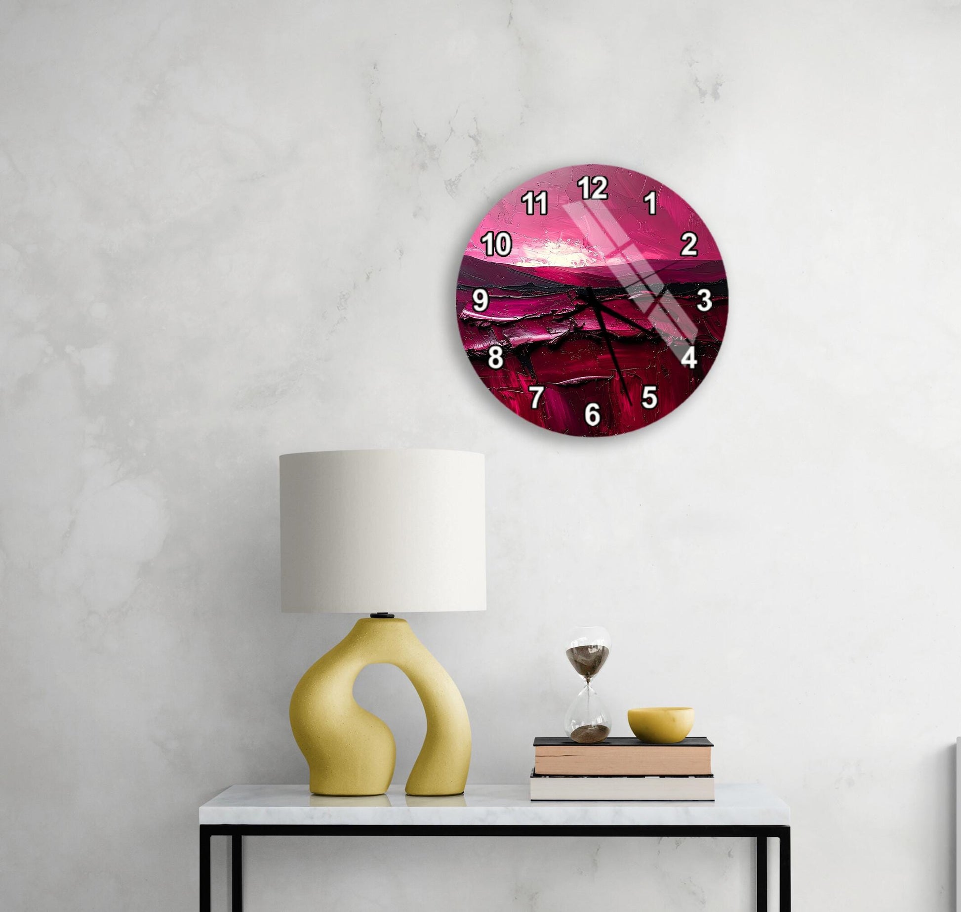 a round wall clock with a pink and purple sunset design, mounted on a white wall above a black table with a yellow lamp and a stack of books.