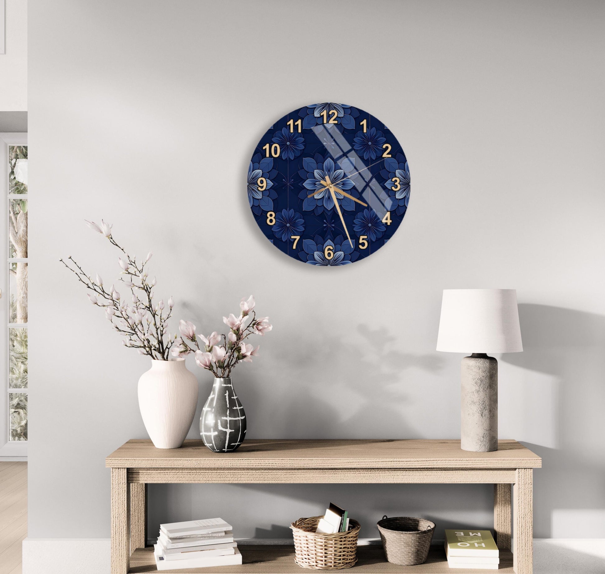 a wall clock with a blue floral design, a wooden table with a lamp, a vase with pink flowers, and a stack of books on a shelf.