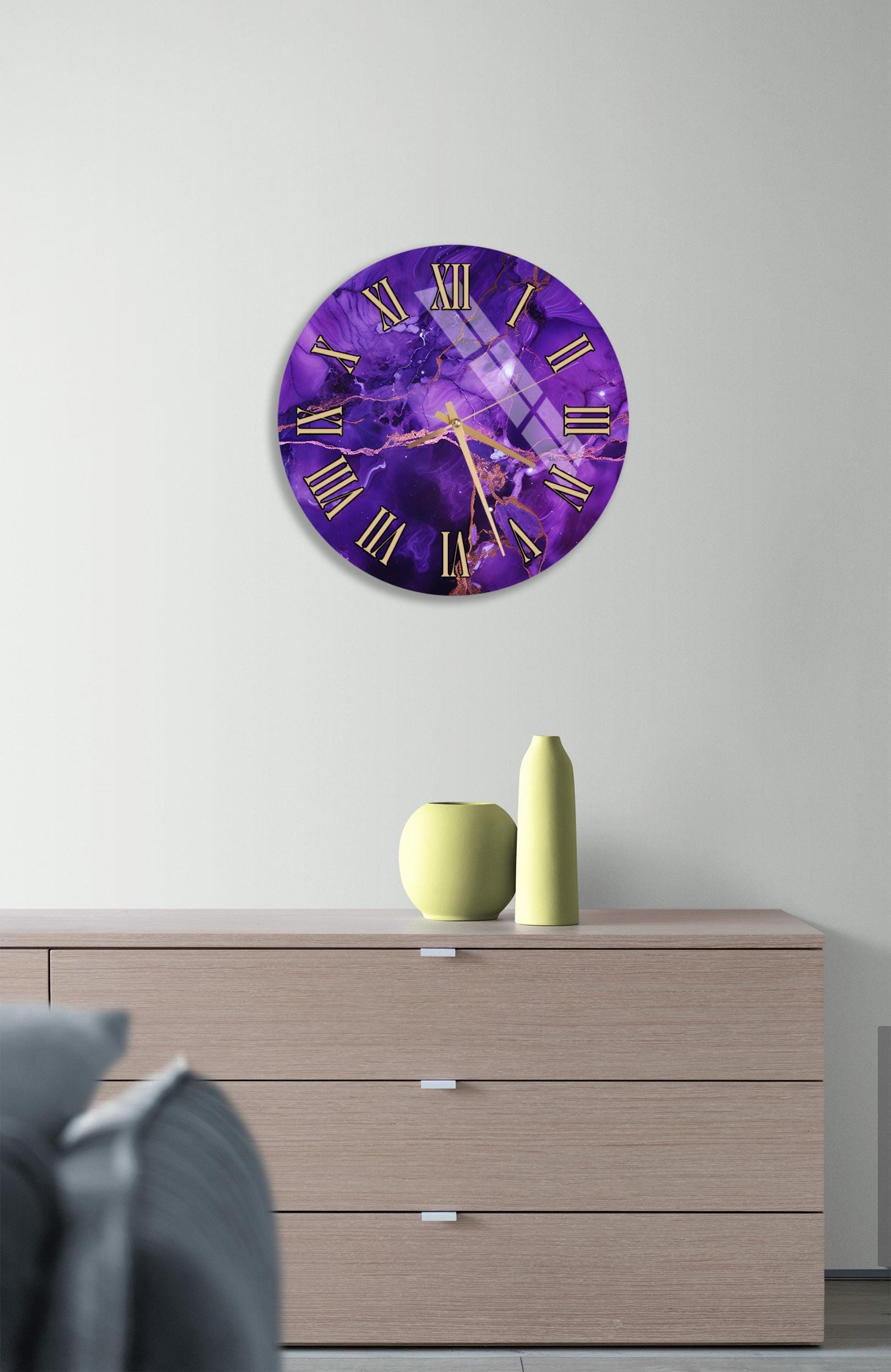 a purple clock with gold Roman numerals, mounted on a wall above a wooden dresser. The dresser has a vase and a small yellow pot on it.