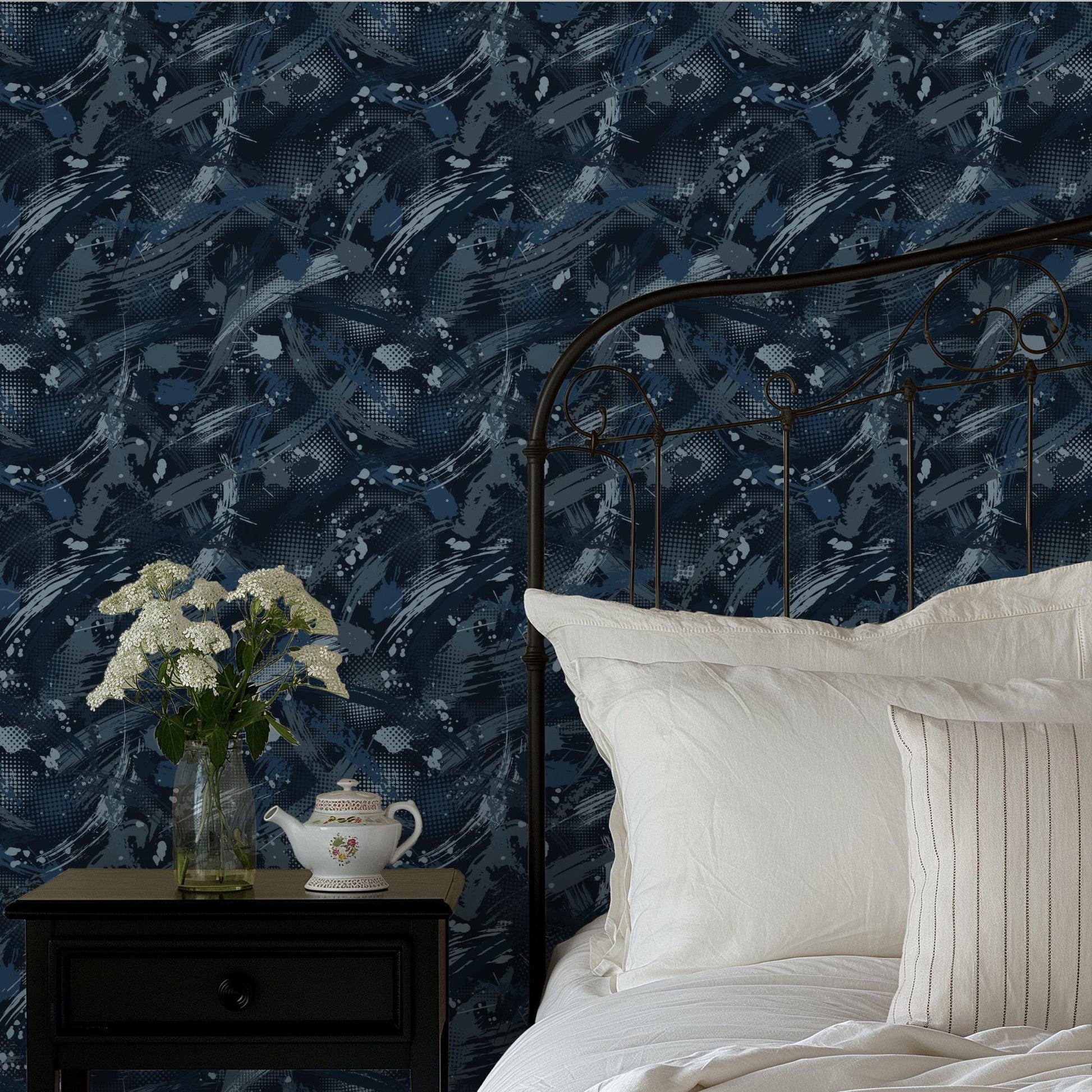 a bedroom with a black metal headboard, a white bedspread, and a wooden nightstand with a teapot and a vase of white flowers. The wall behind the bed has a blue and white abstract pattern.