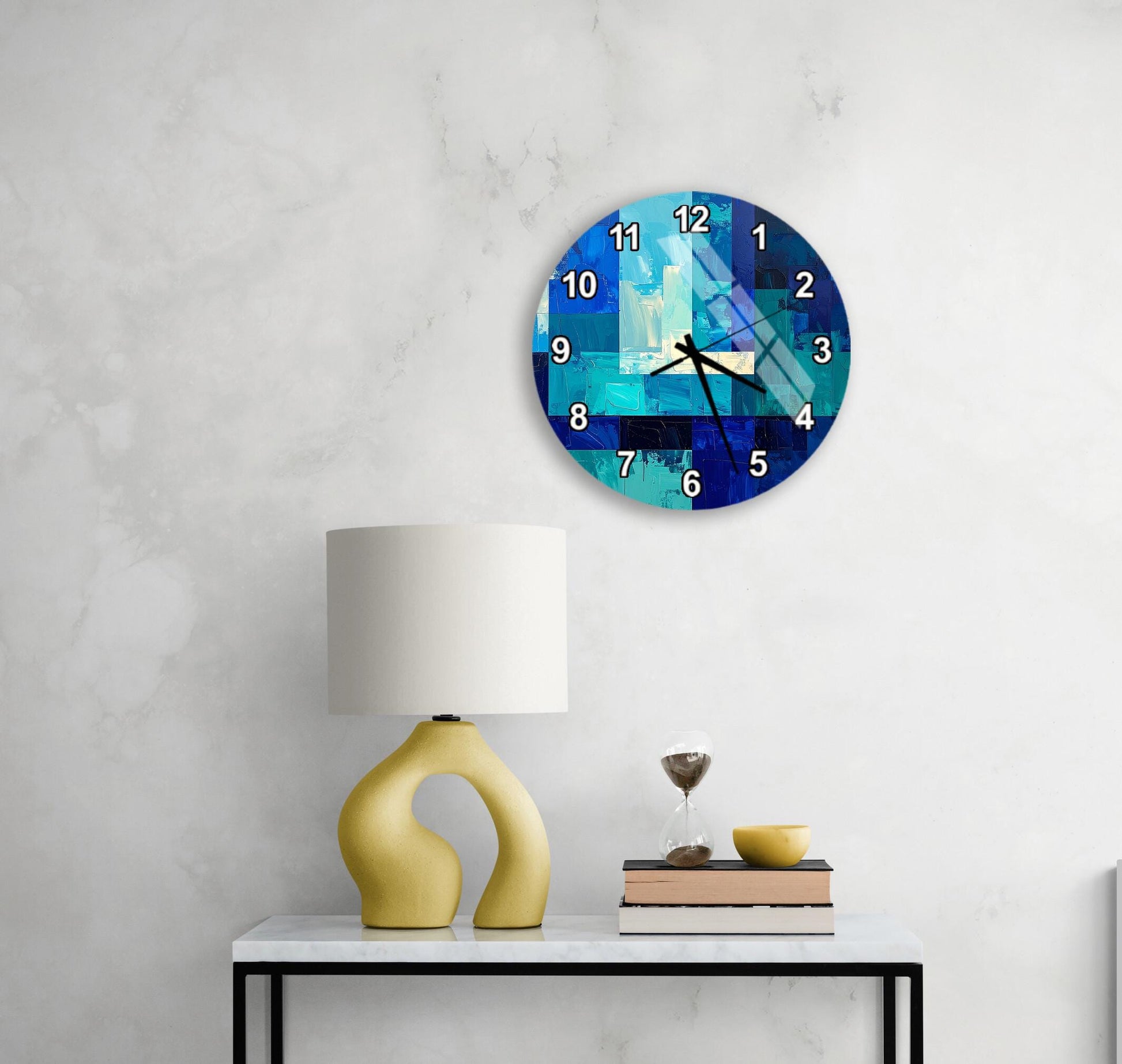a modern wall clock with a blue and white color scheme, mounted on a white wall above a white table with a yellow lamp and a stack of books.