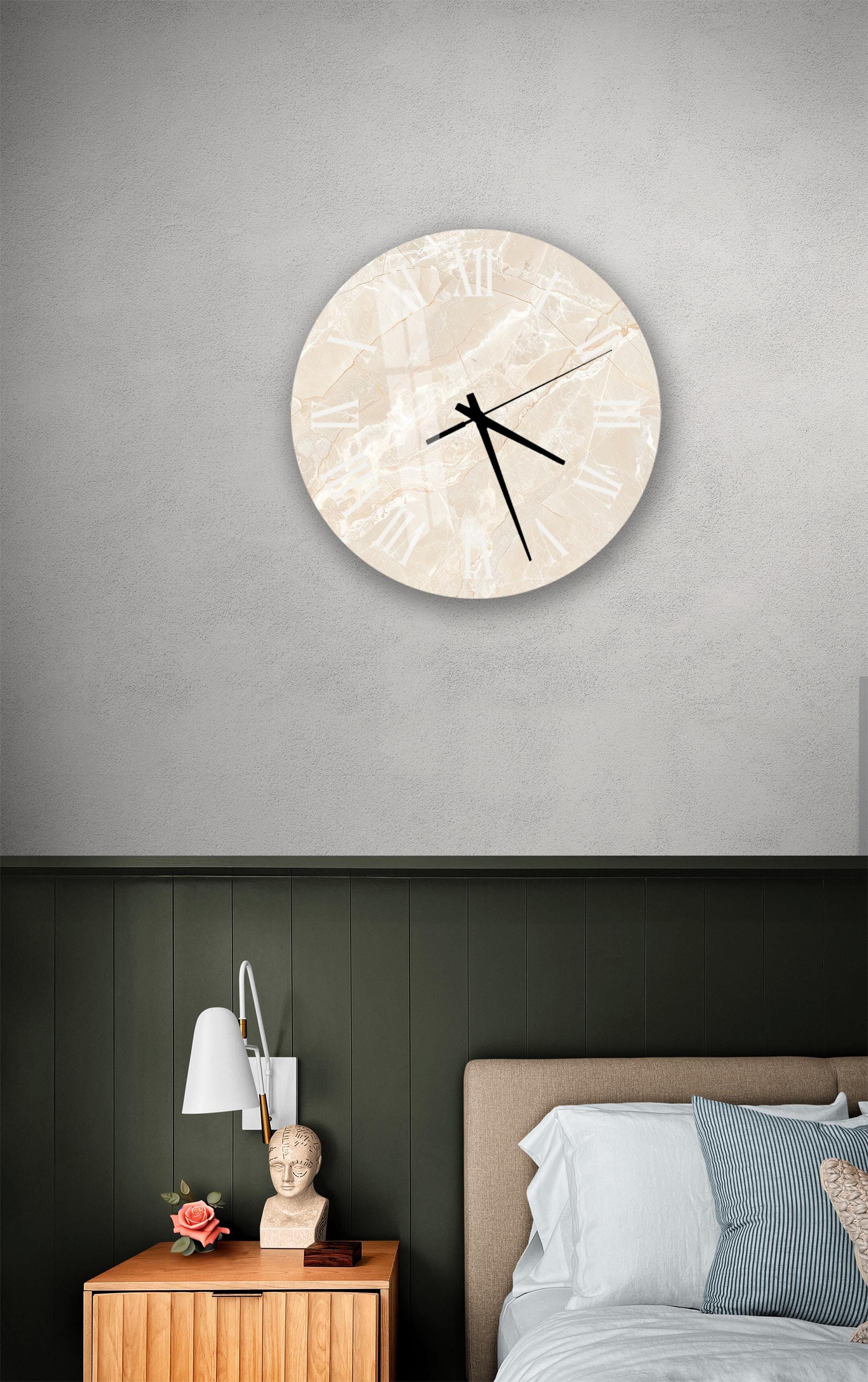 a bedroom setting with a round clock on the wall above a bed. The clock has a marble-like design and is mounted on a gray wall. The bed has a wooden headboard and is covered with white sheets. There is also a nightstand with a lamp and a small statue on it.